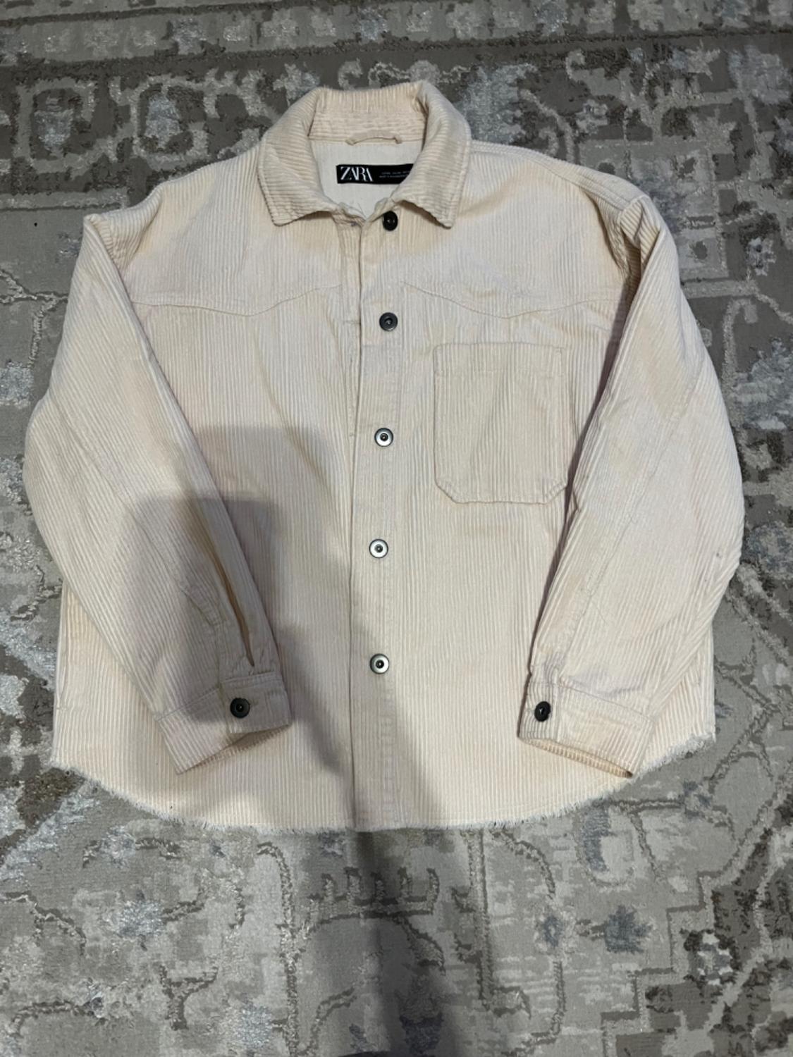 Beige corduroy shirt with black buttons on a gray rug with a geometric design. Visible label on the collar.