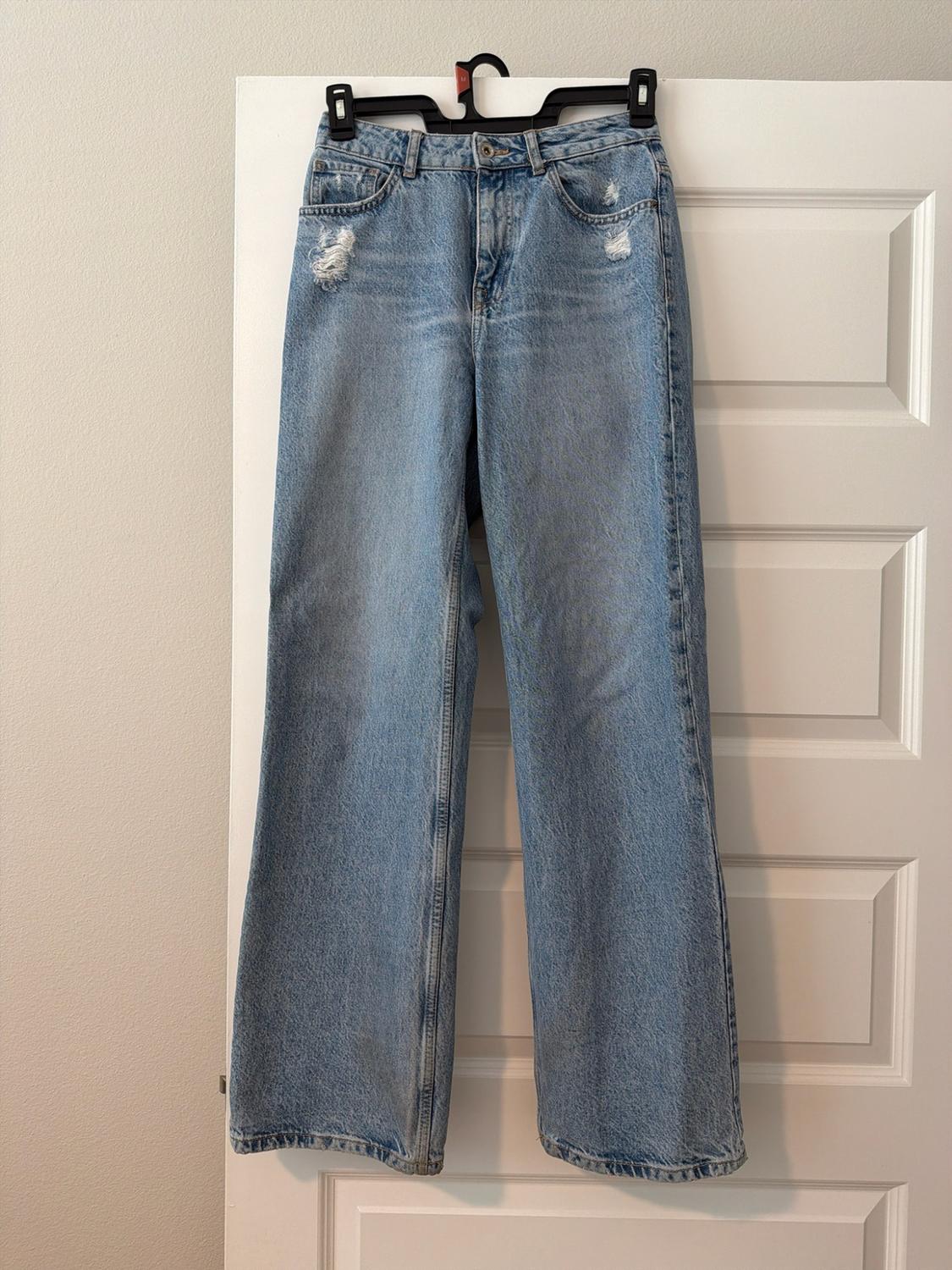 Light blue jeans with ripped detailing below the pockets, hanging on a black hanger over a white door.