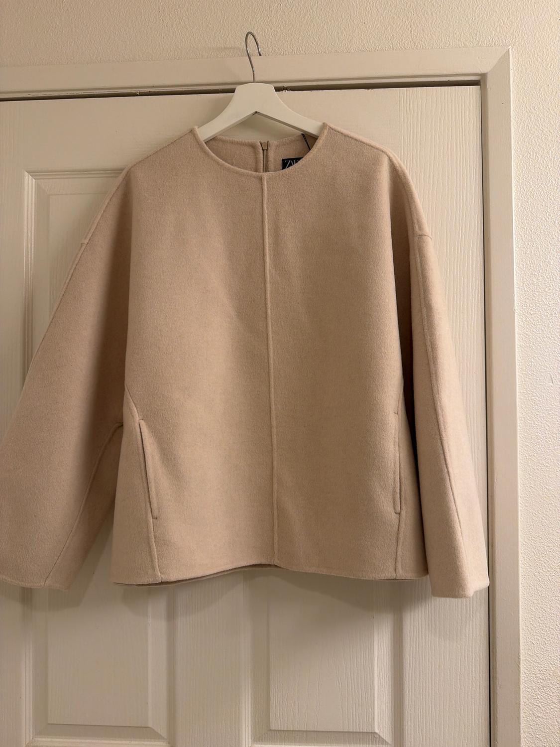 Long-sleeved beige jacket hanging on a white hanger over a door. Minimalist design and straight cut.