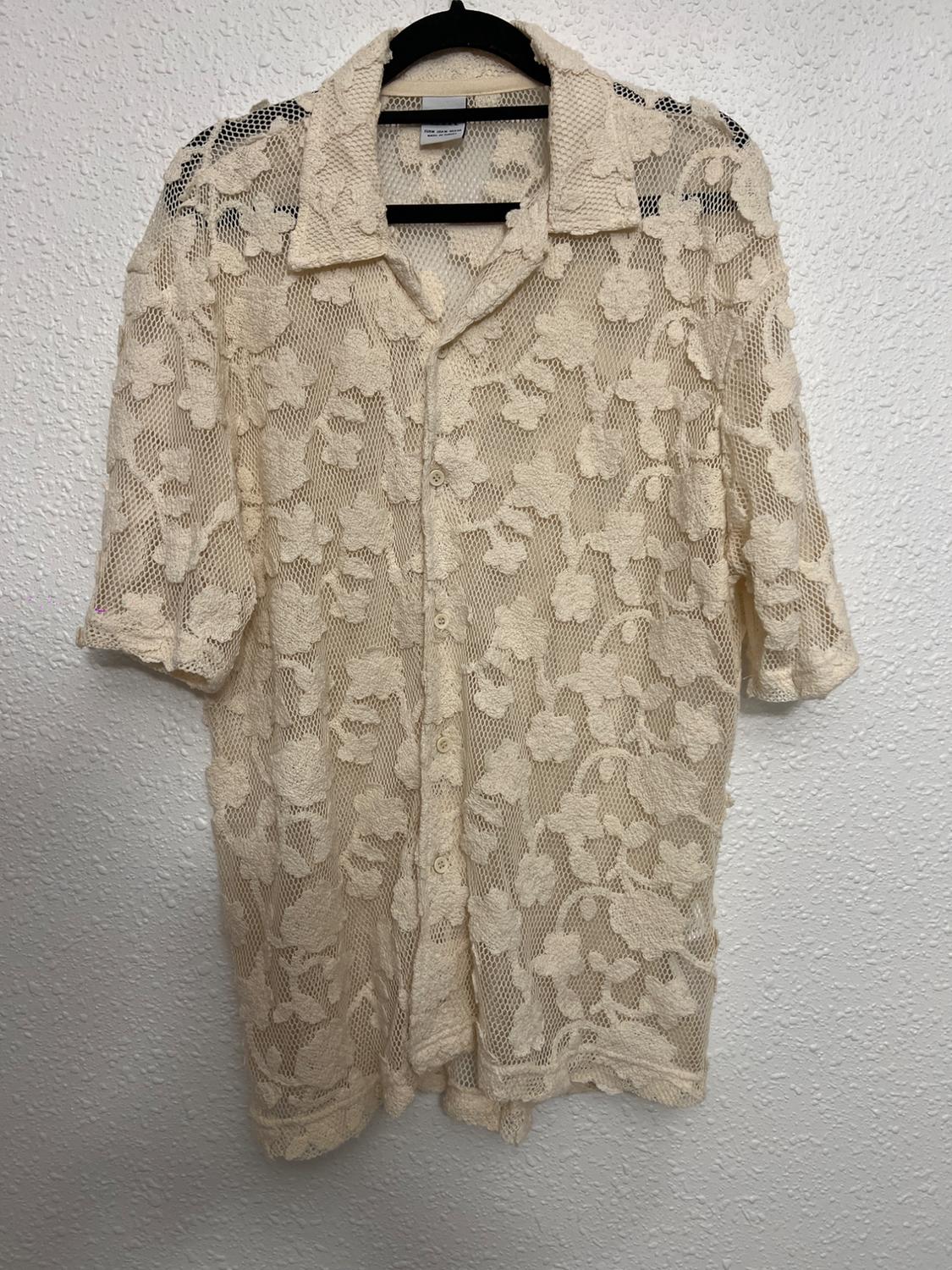 Beige lace shirt with a floral design, hanging on a black hanger against a textured white wall.