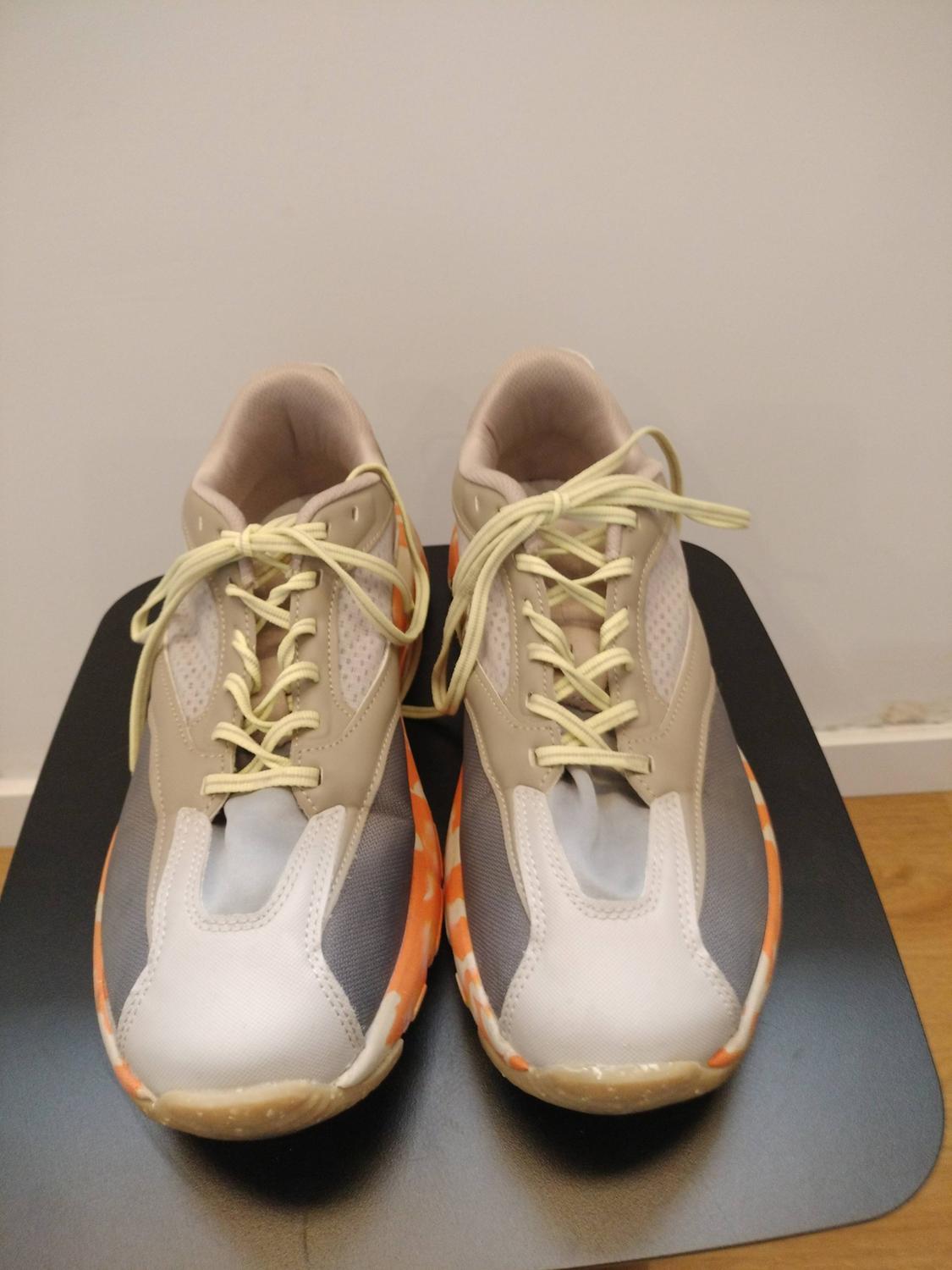 Trainers in beige, grey, and orange tones with yellow laces, on a black surface and white background.