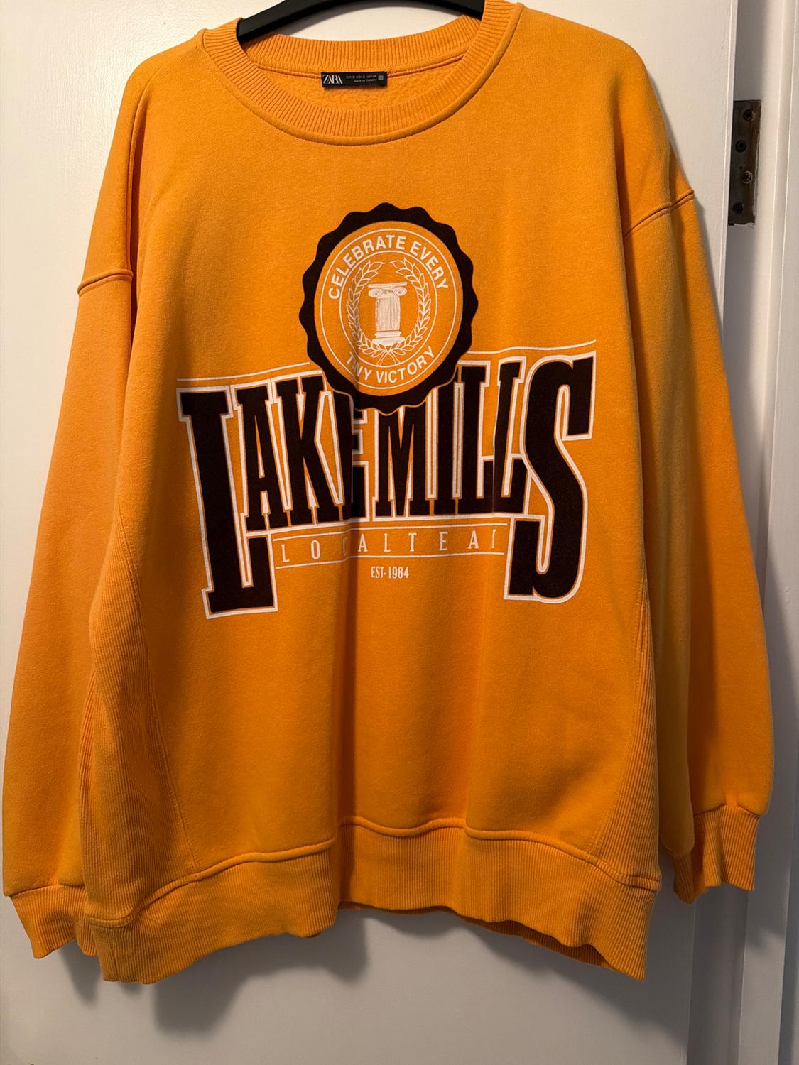 Orange sweatshirt hanging with the text "LAKE MILLS LOCAL TEAM EST. 1984" and "CELEBRATE EVERY VICTORY" in the centre.