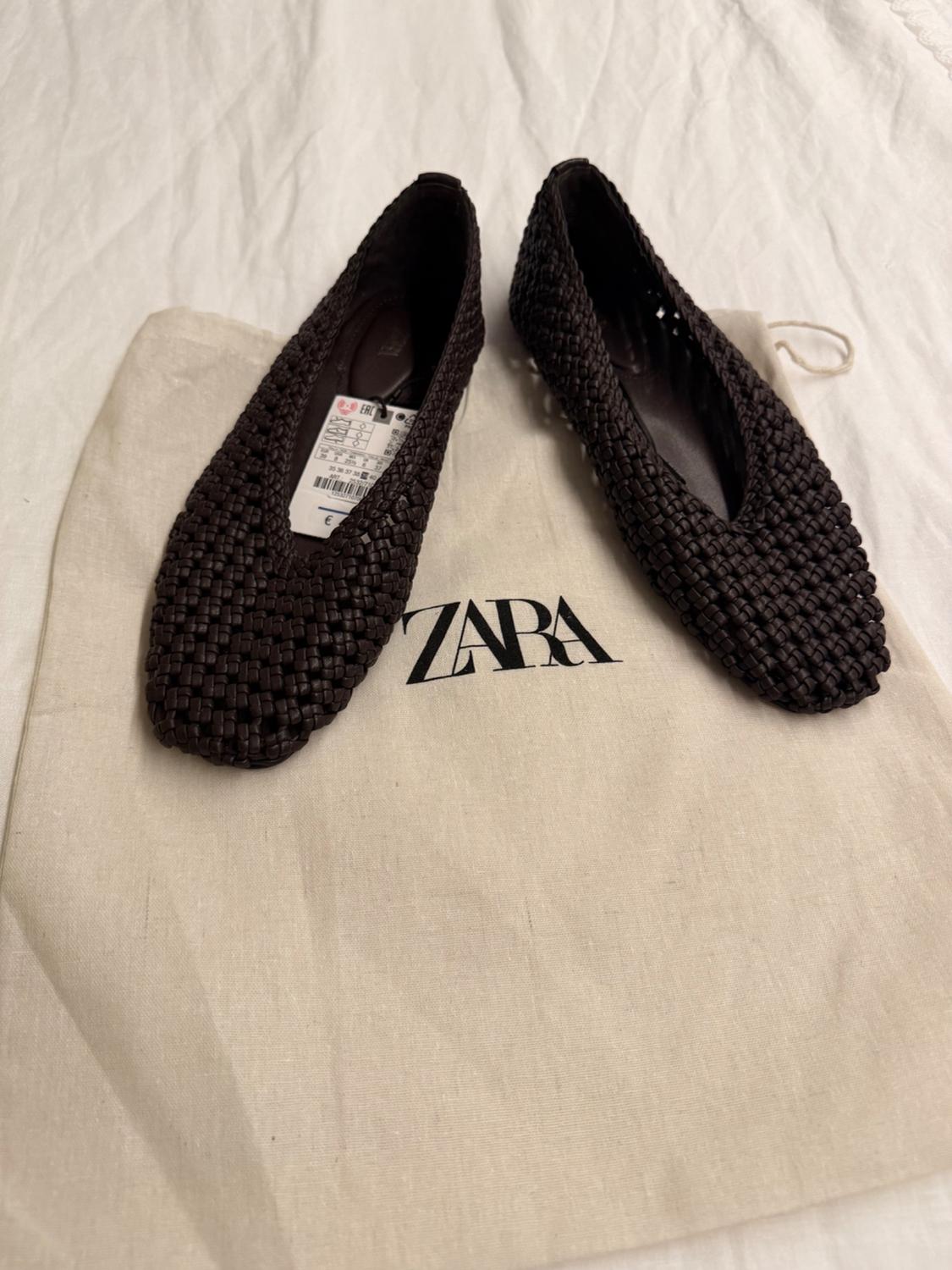 Dark brown braided flat shoes with a tag, on a beige bag with the text ZARA, resting on a white fabric.