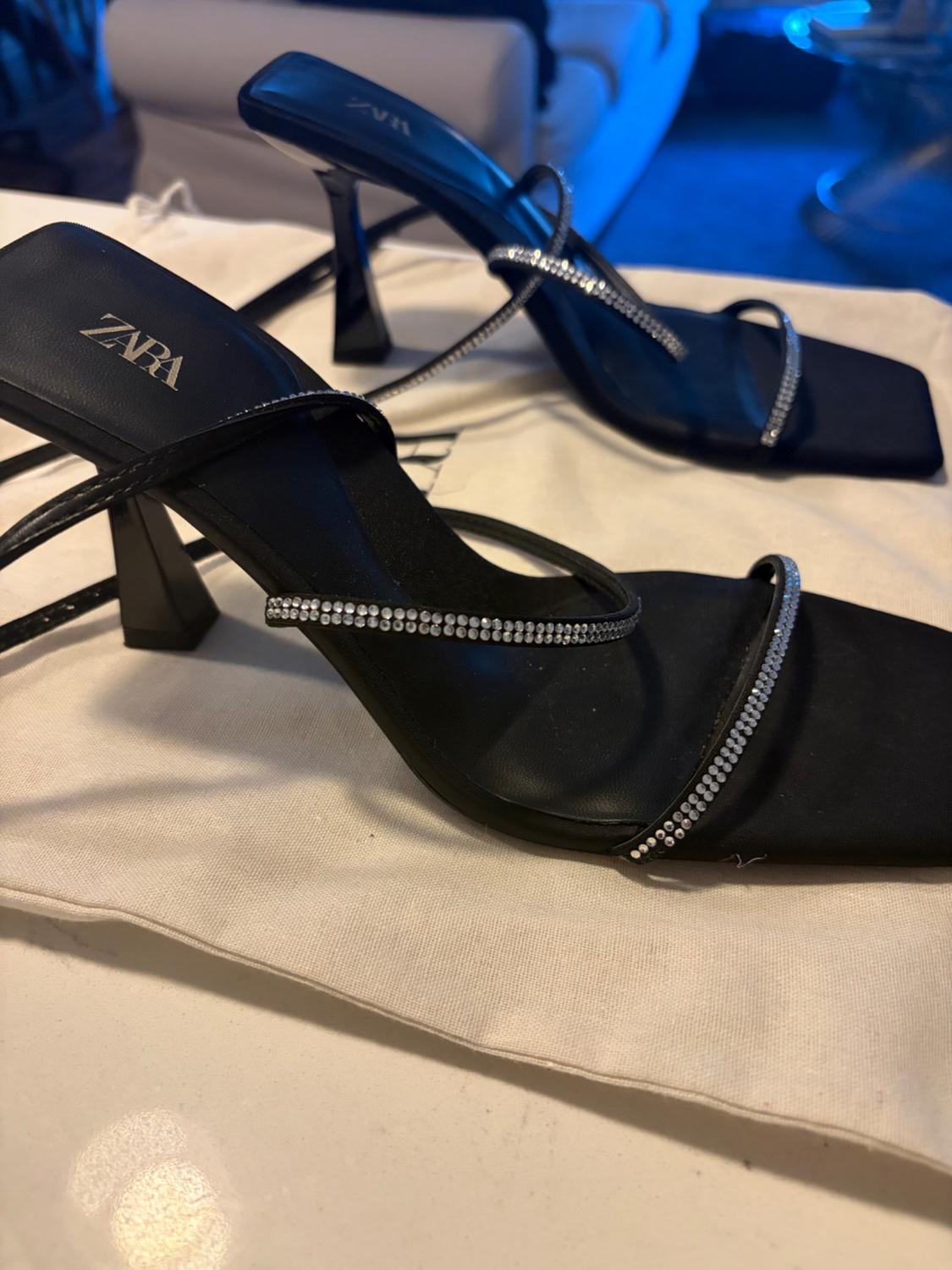 Black heeled sandals with diamante-encrusted straps on a white fabric bag. ZARA text on the insole. Blurred background with blue lighting.