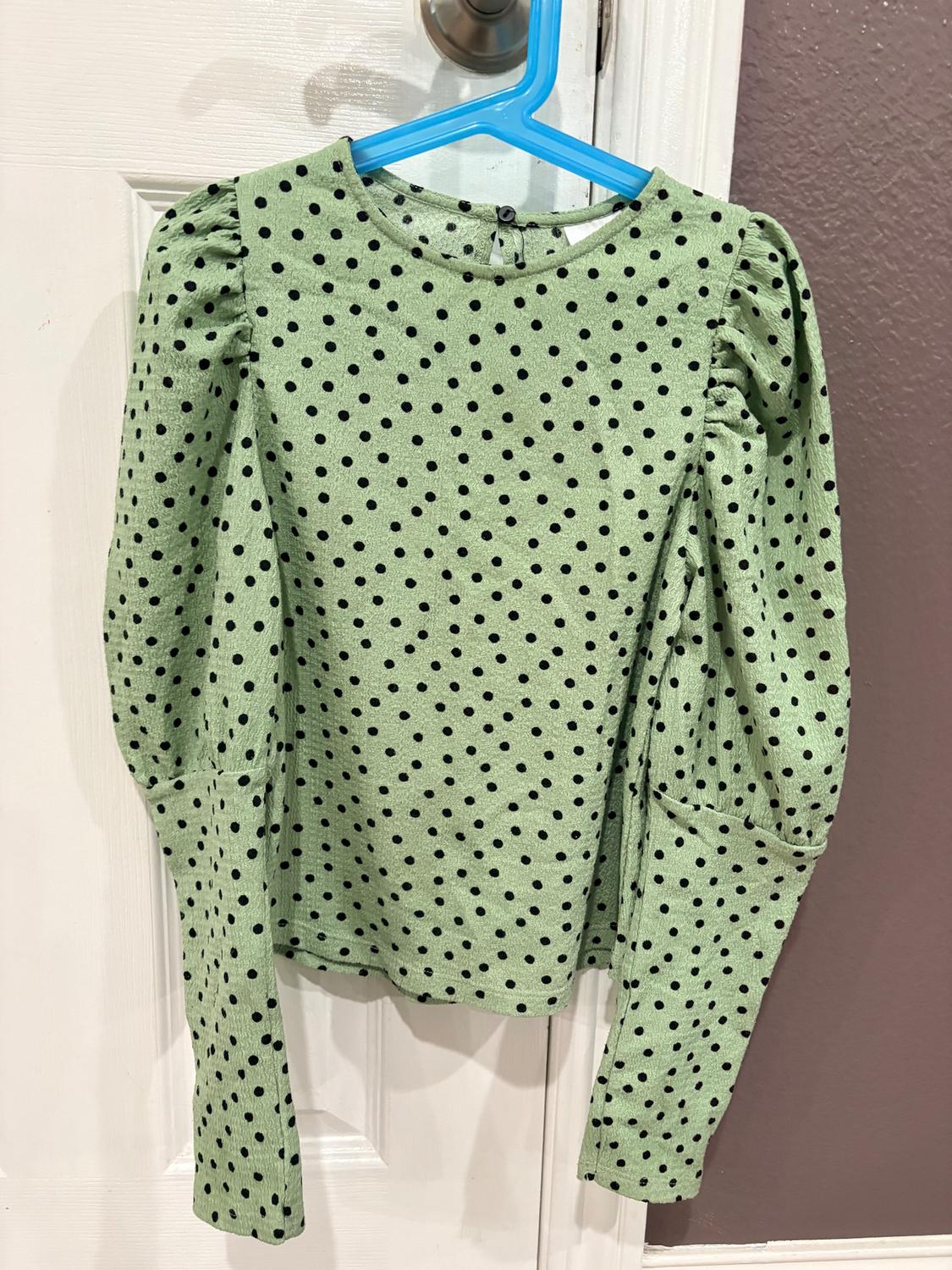 Green blouse with black polka dots and puffed sleeves hanging on a blue hanger over a white door.