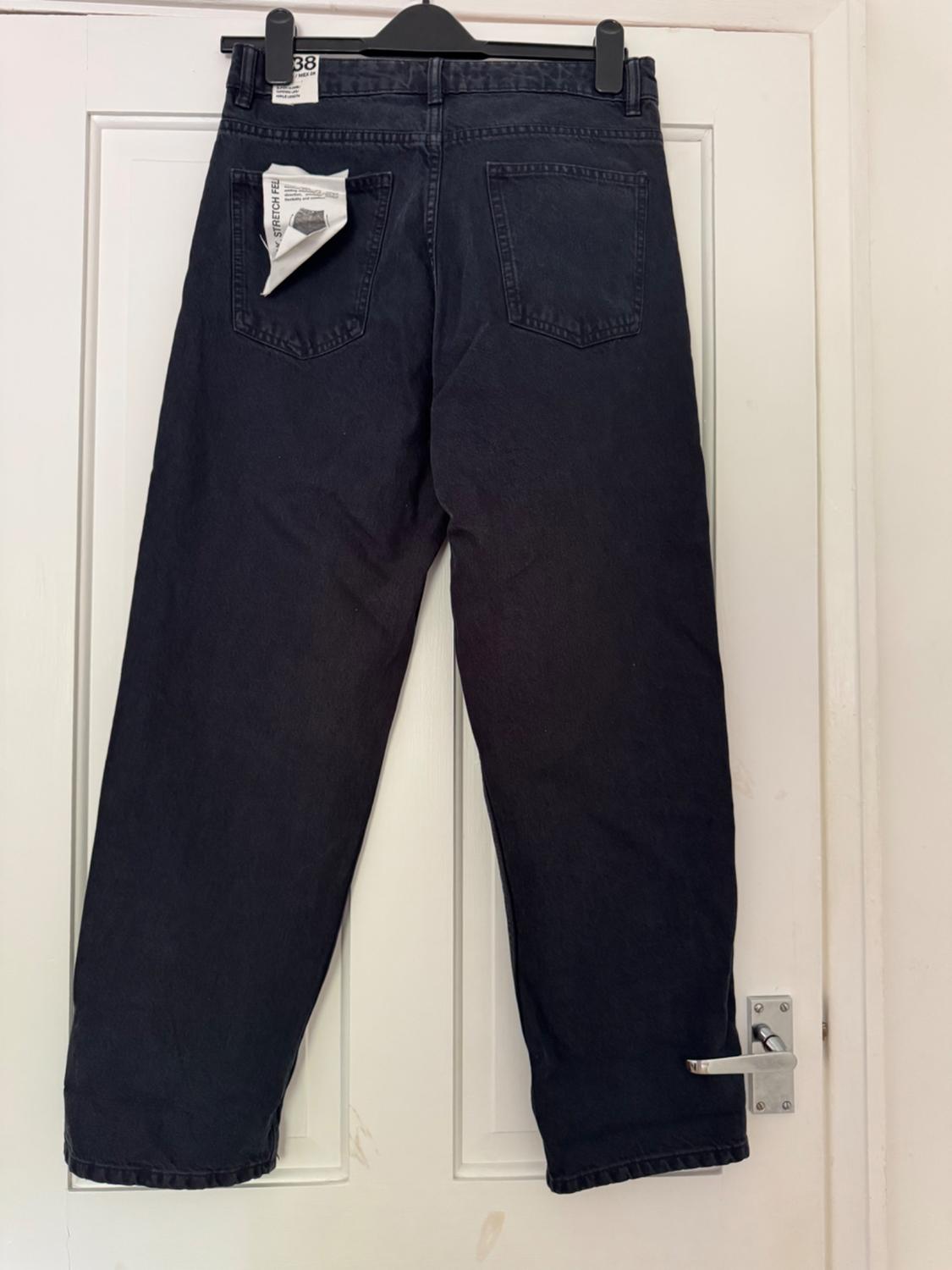 Black jeans seen from behind, hanging on a hanger on a white door. Includes labels with the text 38 and STRETCH.