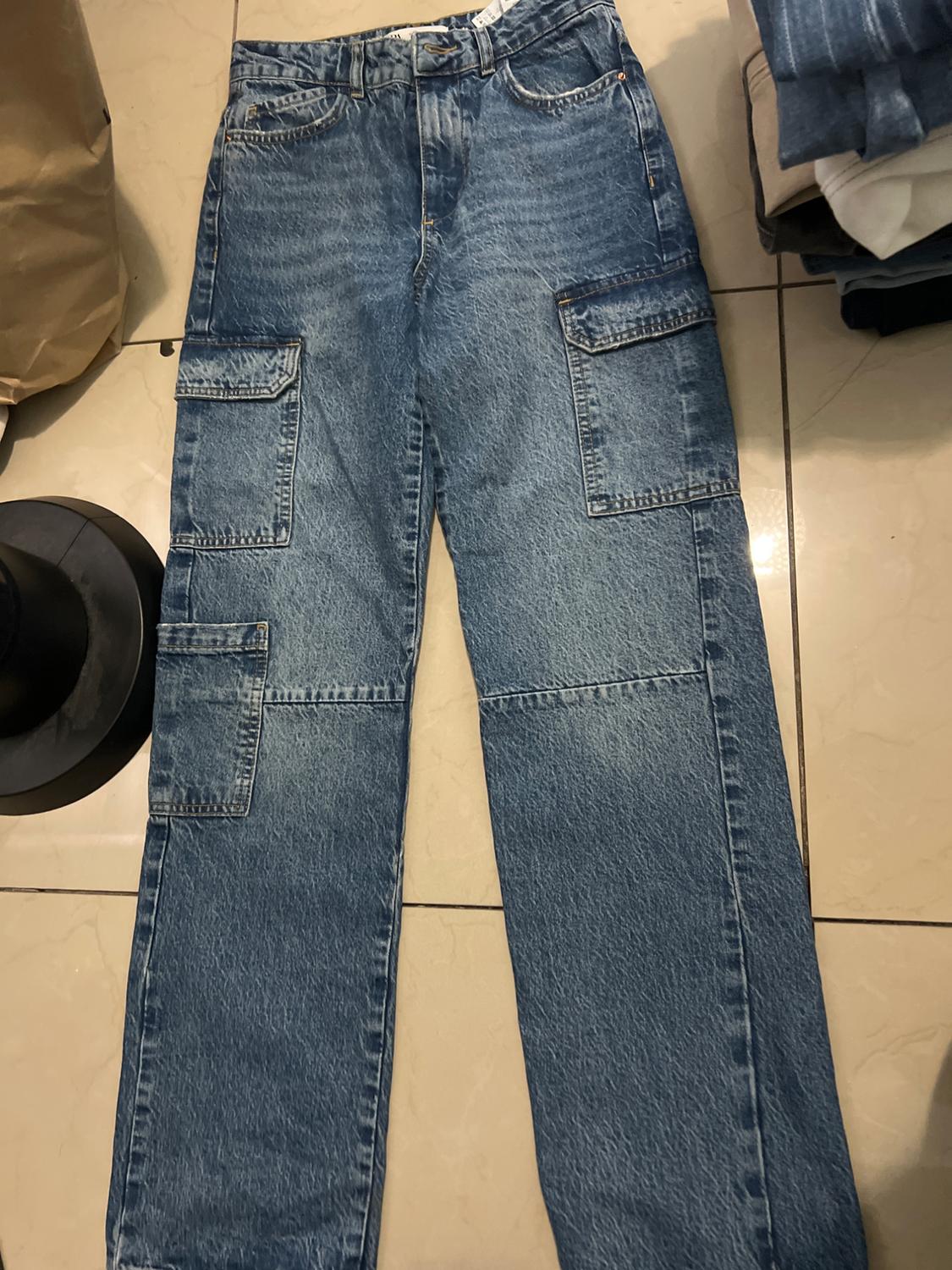 Blue denim cargo trousers with multiple pockets, placed on a tiled floor next to a black hat and folded clothing.