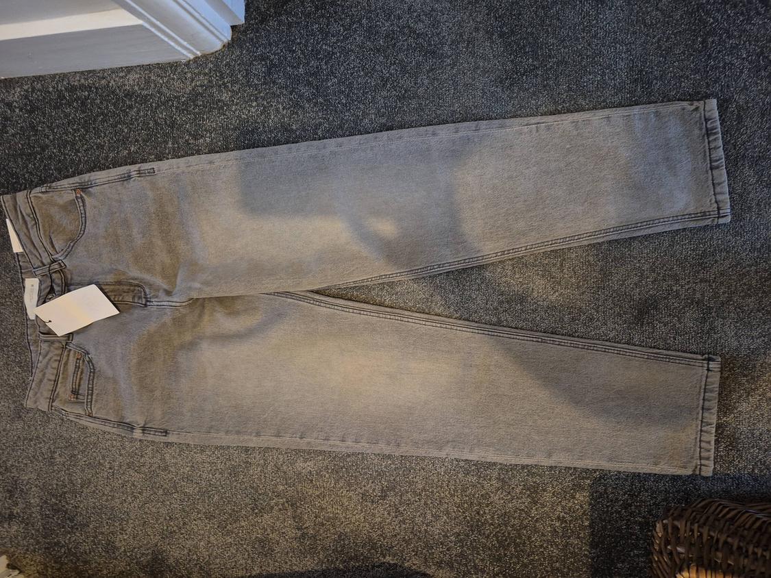 Grey denim jeans laid out on a grey rug. White tags visible on the left front pocket.