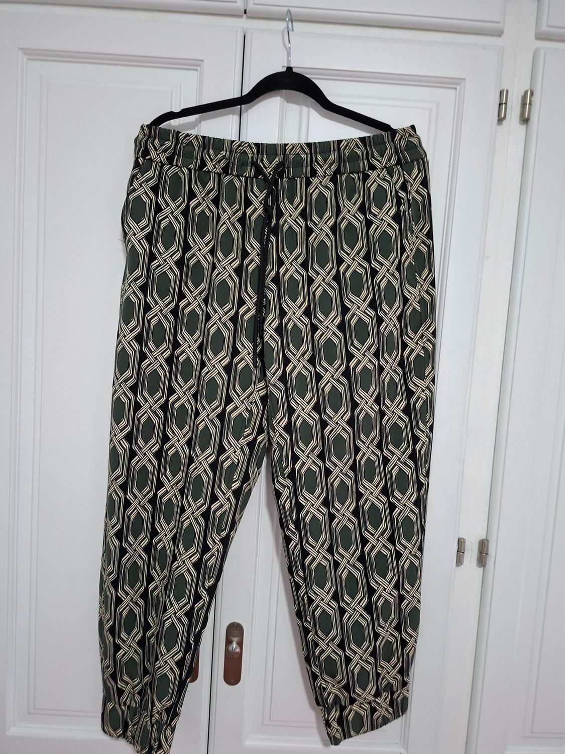 Trousers with a geometric print in green and beige tones, hanging on a black hanger in front of a white wardrobe.