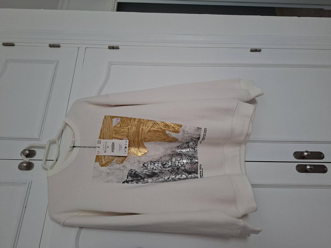 Cream sweatshirt with a gold brushstroke and rocky landscape print, hanging in a white wardrobe. Text on garment: martre 1974.