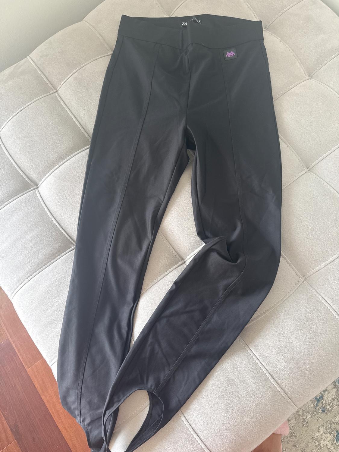 Plain black fabric trousers with an elasticated waist, placed on a padded beige sofa. Small label visible at the top.