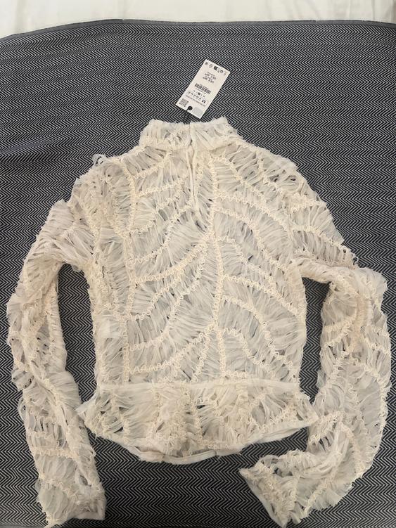 White lace blouse with a leaf design on grey herringbone fabric. Visible tag with price and size details.