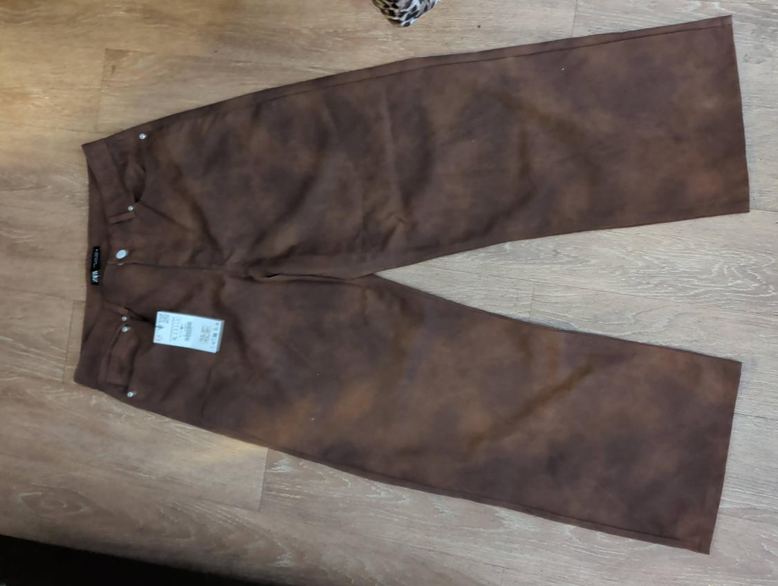 Brown straight-leg pants on a wooden floor. Visible white tag with price and size details.