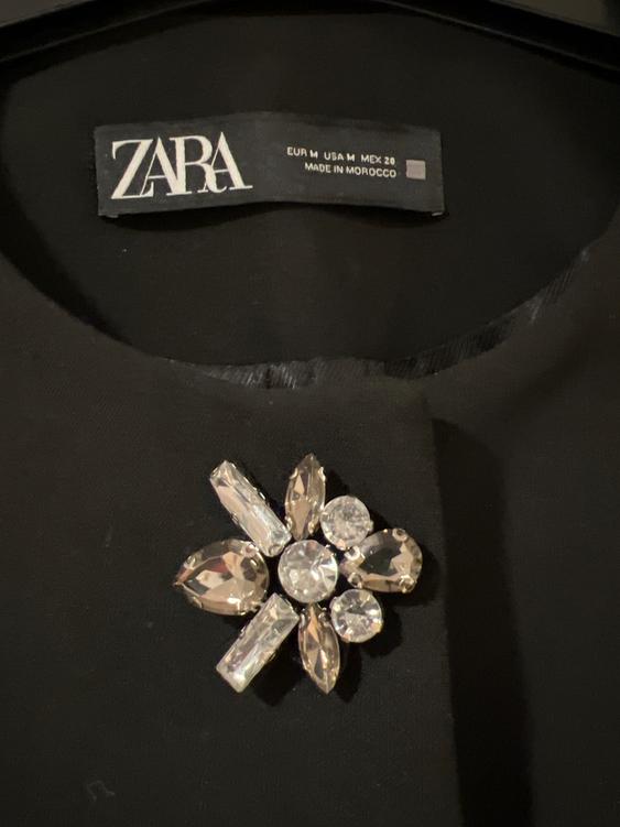 Zara label on a black garment with a rhinestone flower-shaped brooch. Visible text: "EUR M USA M MEX 28 MADE IN MOROCCO".