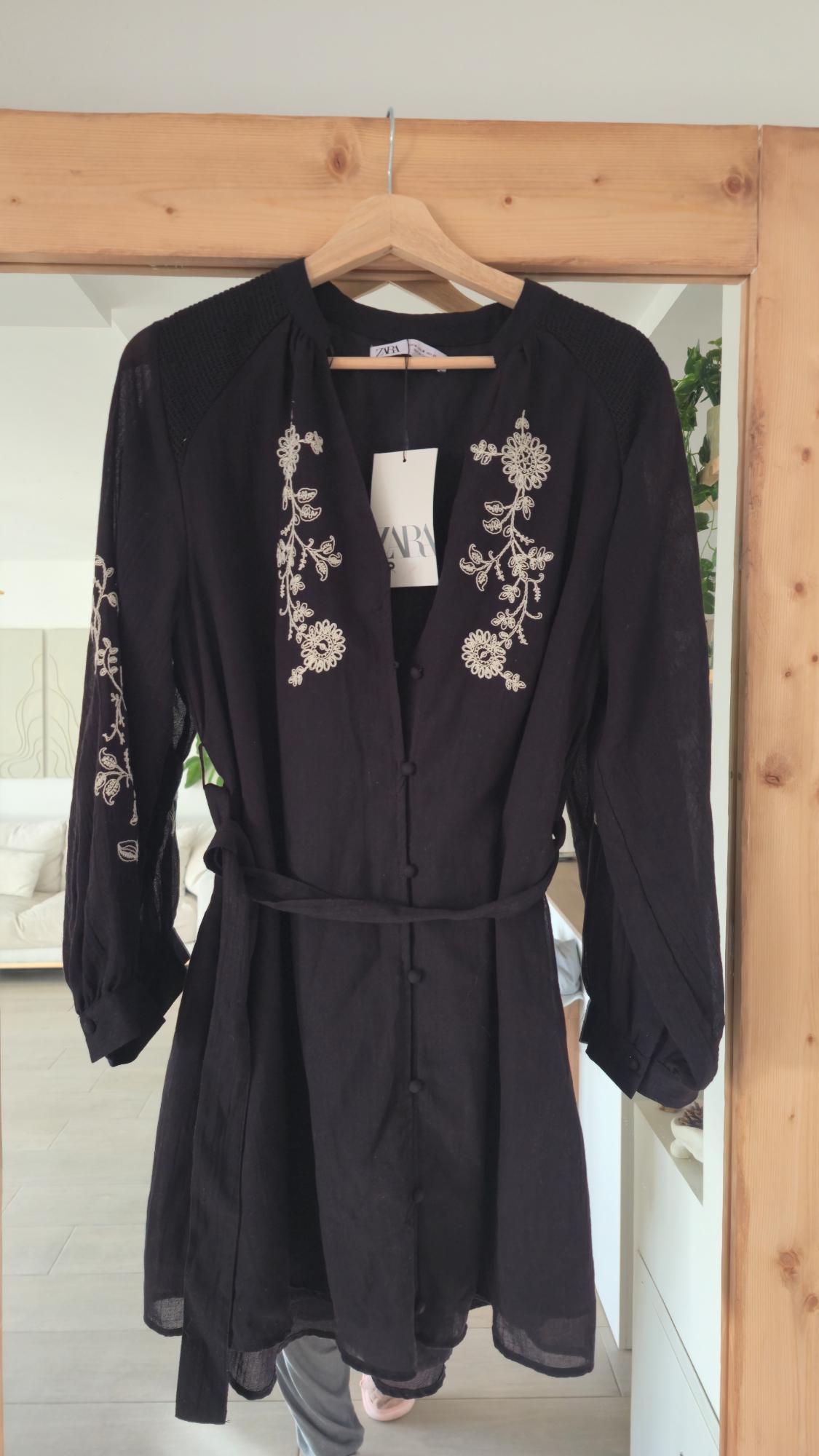Black blouse with white floral embroidery hanging on a wooden hanger. Visible label with the text "ZARA". Modern living room background.