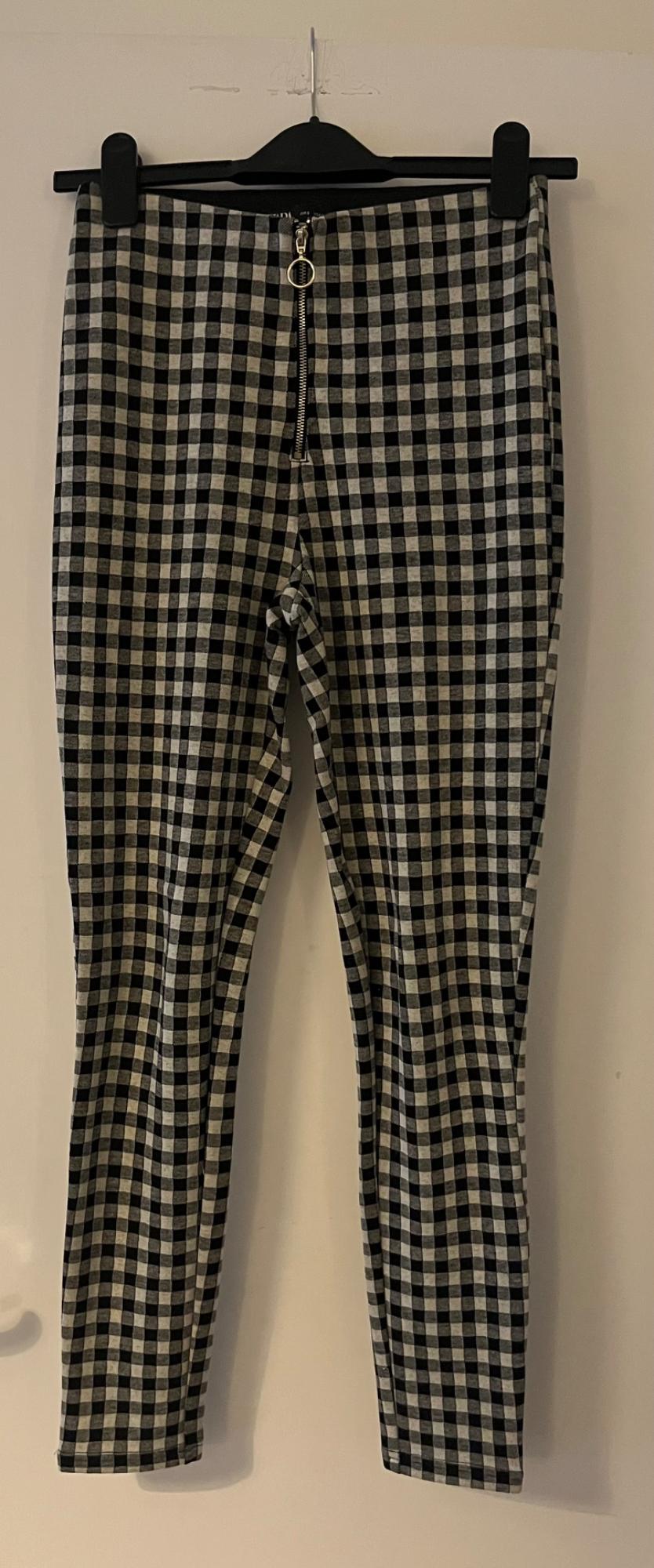 Black and white checked skinny trousers on a black hanger against a white background. Featuring a front zip with a ring pull.