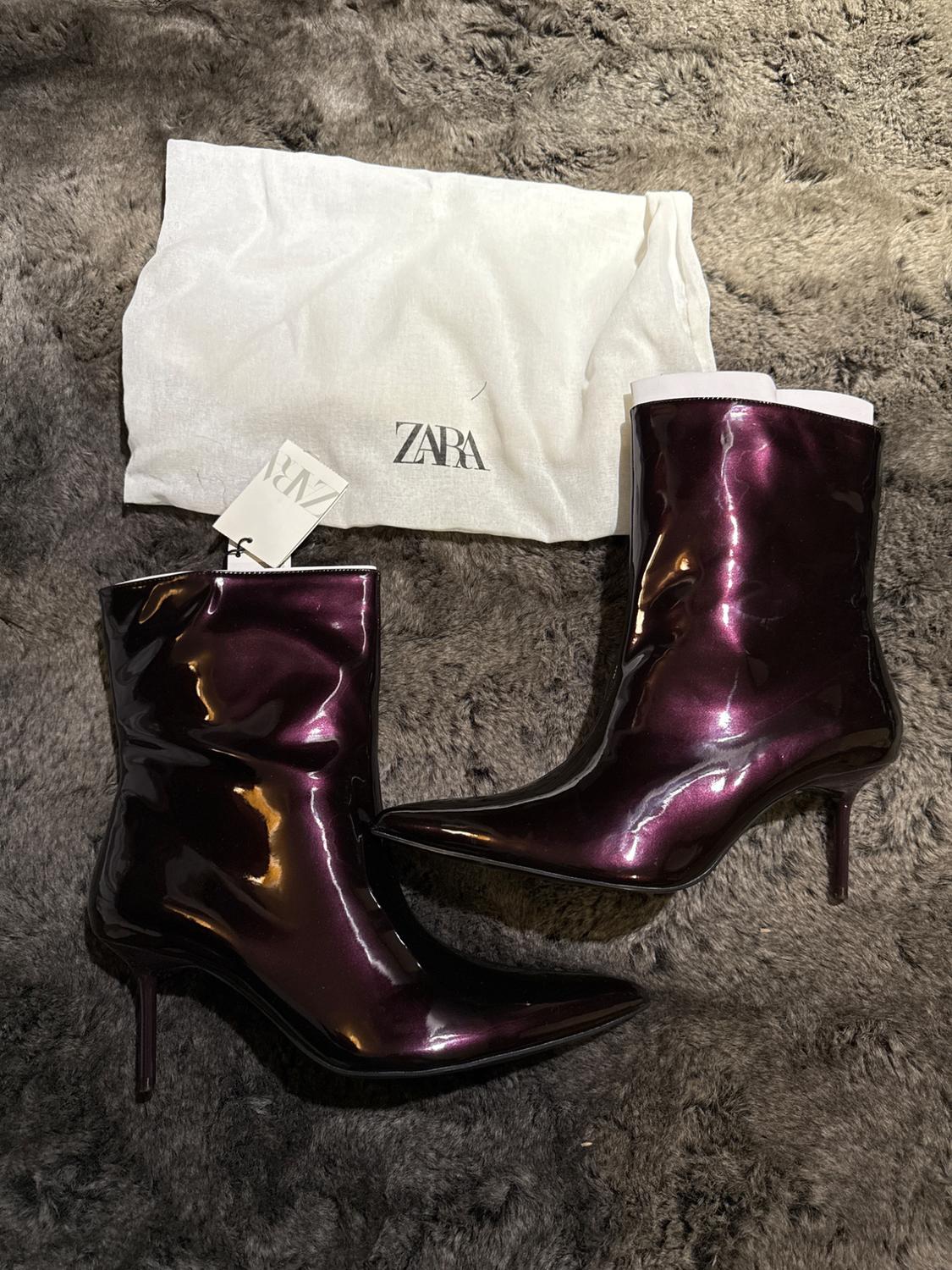 Dark purple patent leather ankle boots with stiletto heels on a gray carpet. White bag with "ZARA" logo and visible tag.