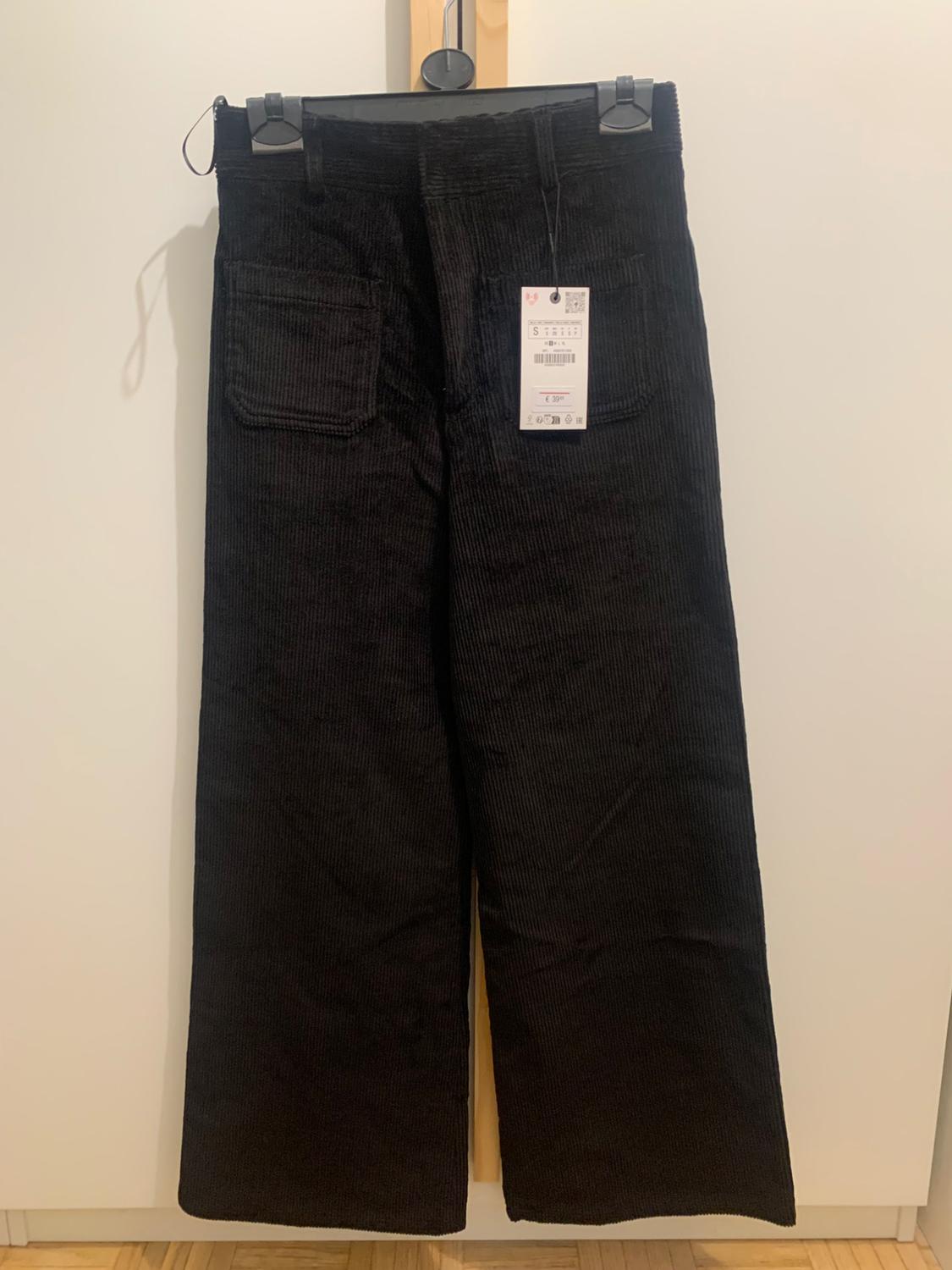 Black corduroy trousers hanging on a hanger with a visible tag. White background and light wooden floor.