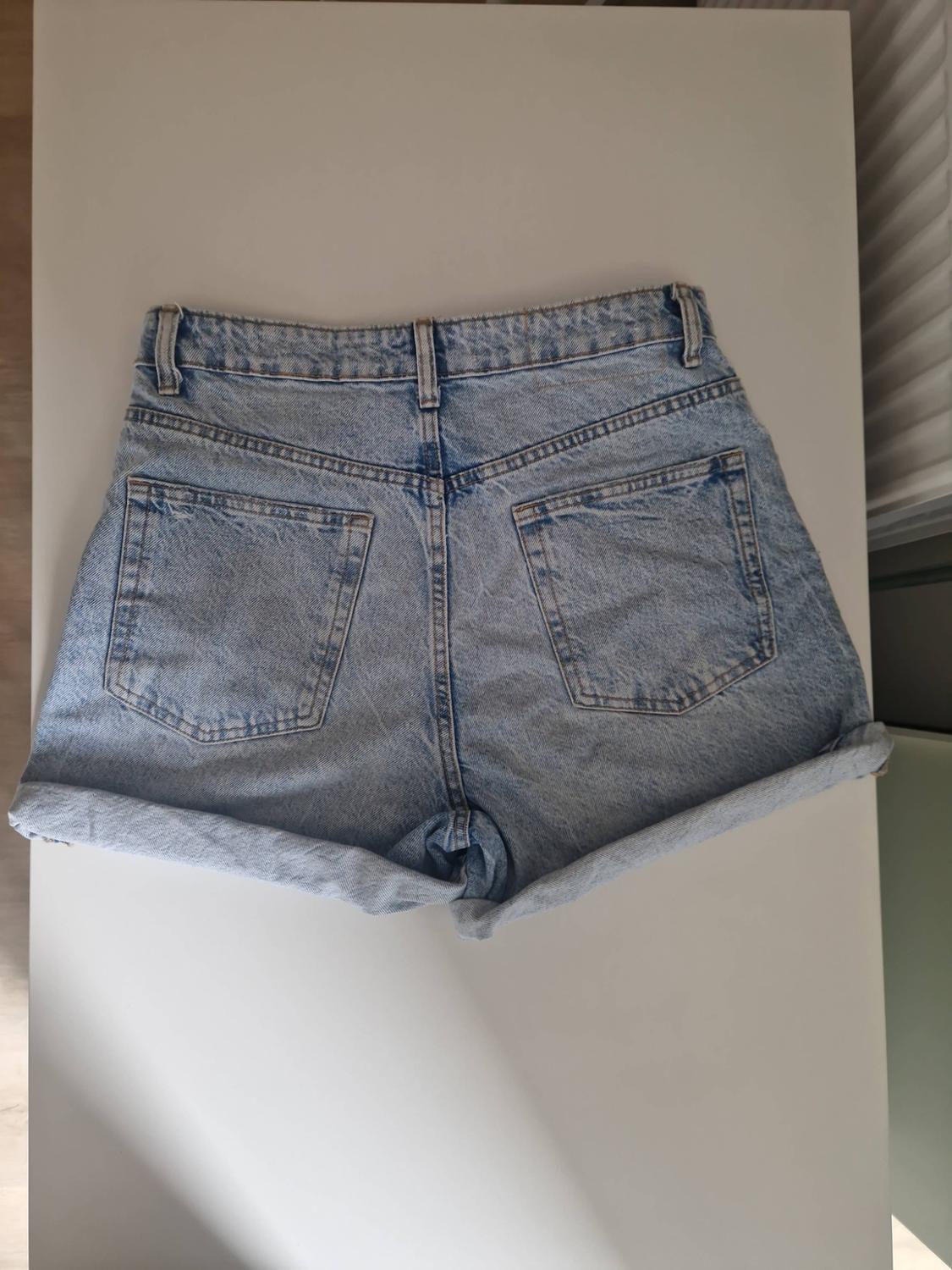 Light blue denim shorts seen from behind, with rolled-up hems and pockets, on a white table with a diagonal shadow.