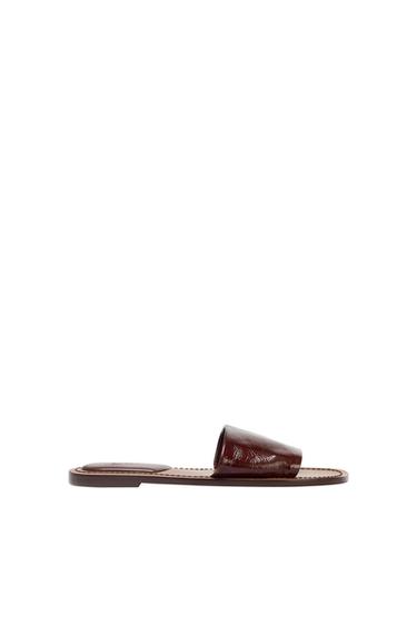 Brown flat sandal, side profile, on a white background. Minimalist design with a wide strap across the front.