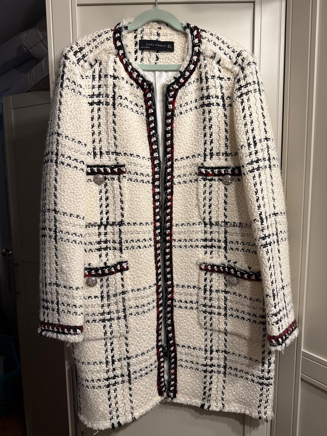White coat with black plaid print and red details, hanging on a green hanger. Visible tag: "ZARA WOMAN".