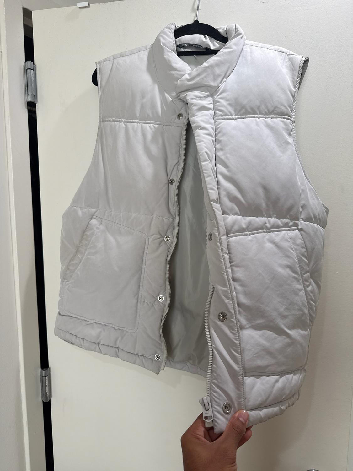 A light gray quilted vest, featuring a zipper and buttons, hangs from a white door. A hand holds the bottom hem of the garment, which is partially open.