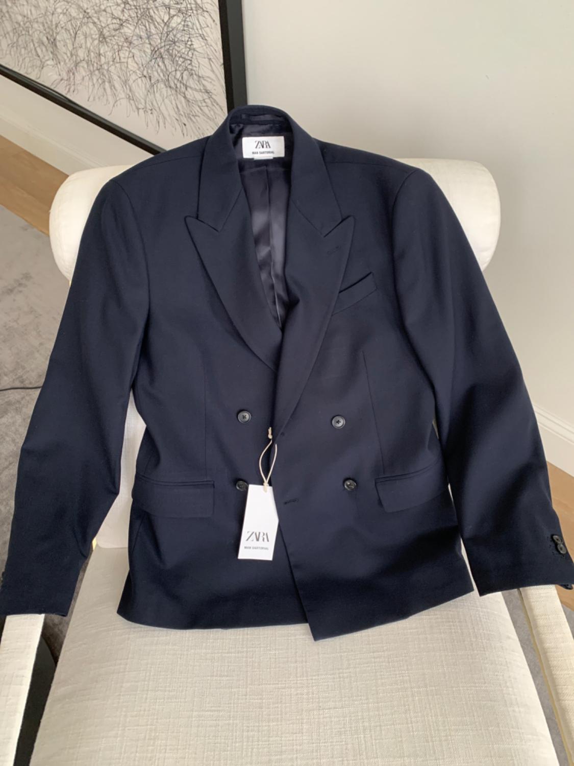 Navy double-breasted jacket on a white chair, with "ZARA" label visible. Background with abstract art.