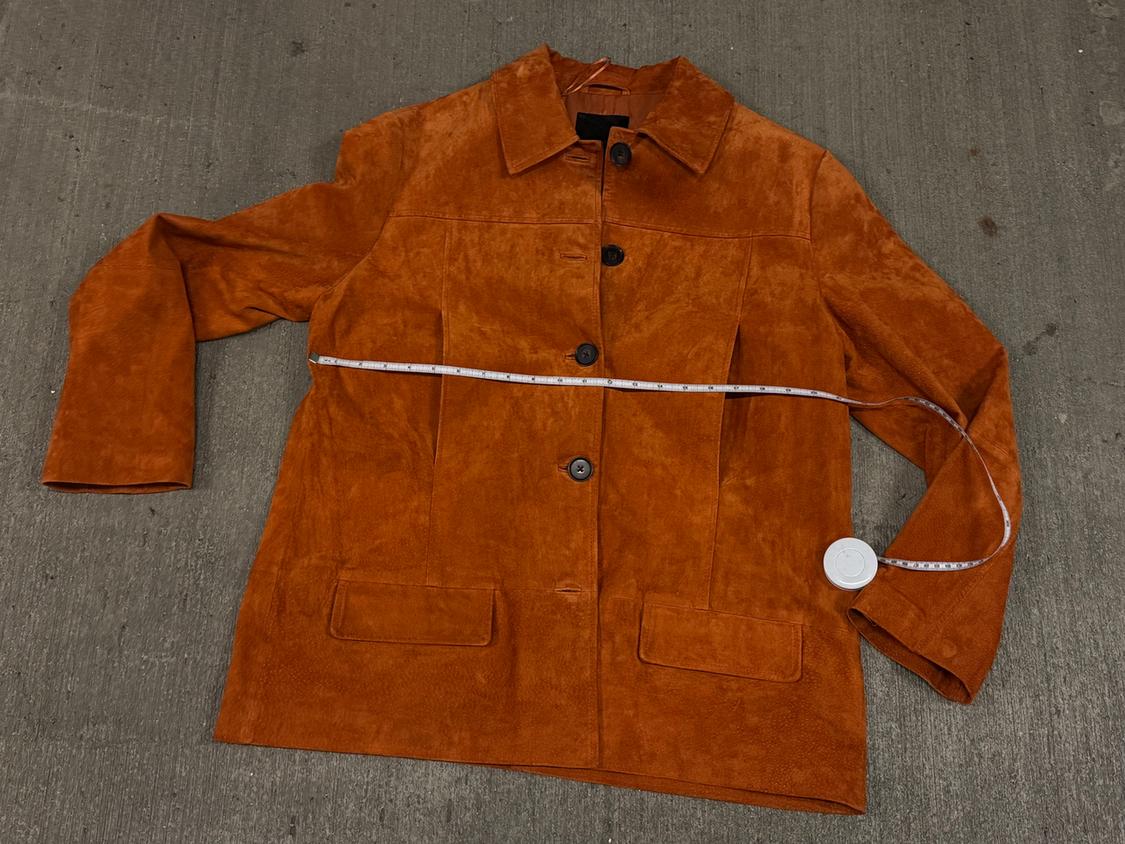 Orange-brown suede jacket with black buttons, placed on a gray surface. White measuring tape around the chest.