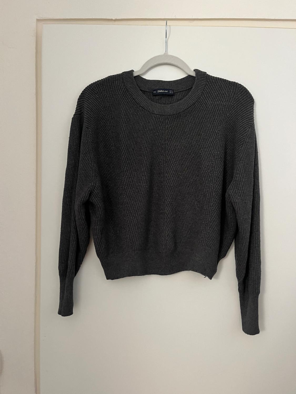 Dark grey knitted jumper hanging on a white hanger over a white door. Visible label with text "ZARA".
