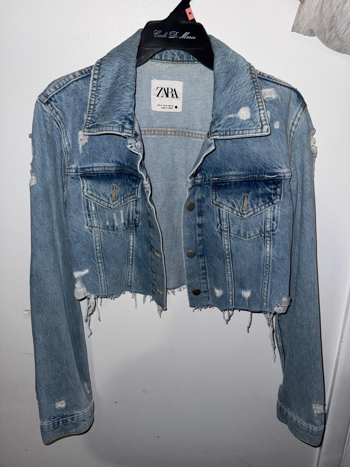 Light blue cropped distressed denim jacket hanging on a black hanger against a white background. Text: Carlo De Marco, ZARA.