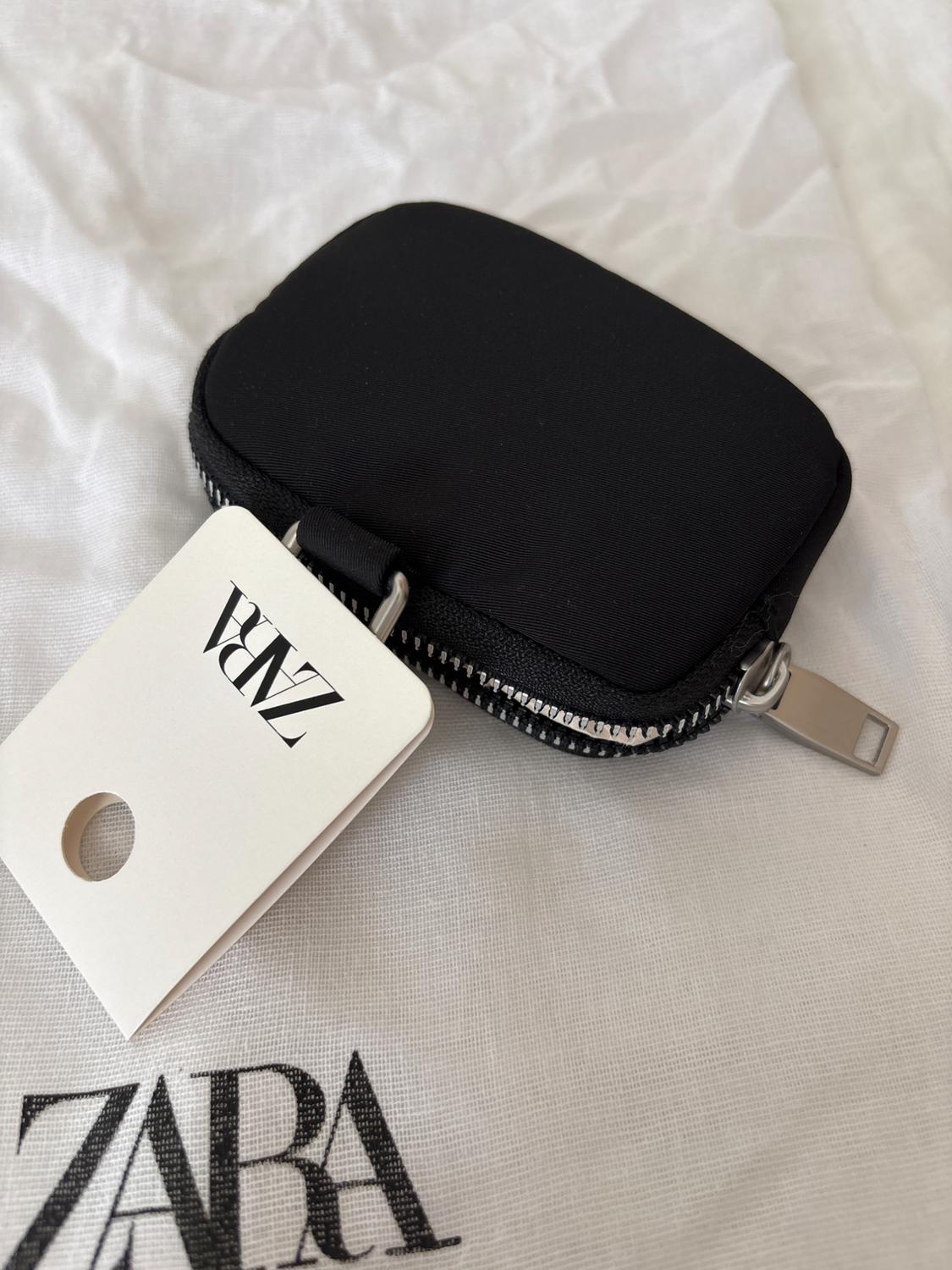Black zippered coin purse on white fabric. Label with text "ZARA" visible. Minimalist and elegant atmosphere.