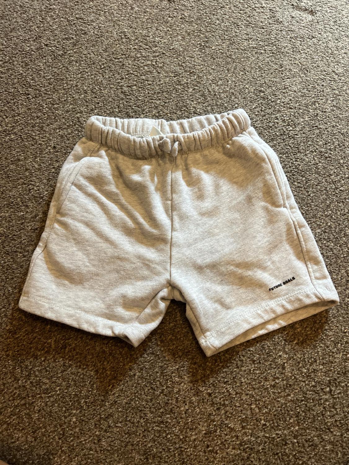 Grey casual shorts with an elasticated waistband on a textured rug. "FUTURE GOALS" text detail on the hem.
