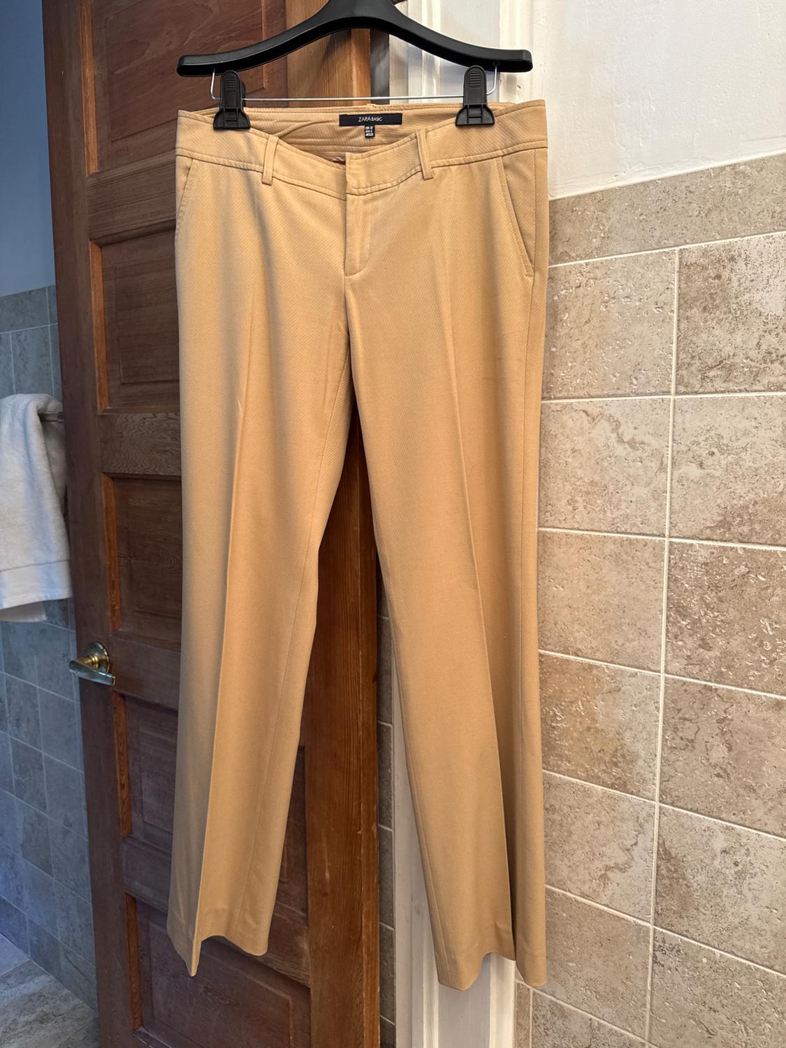 Beige pants hanging on a black hanger over a wooden door, with a beige tiled background and a white towel on the left.