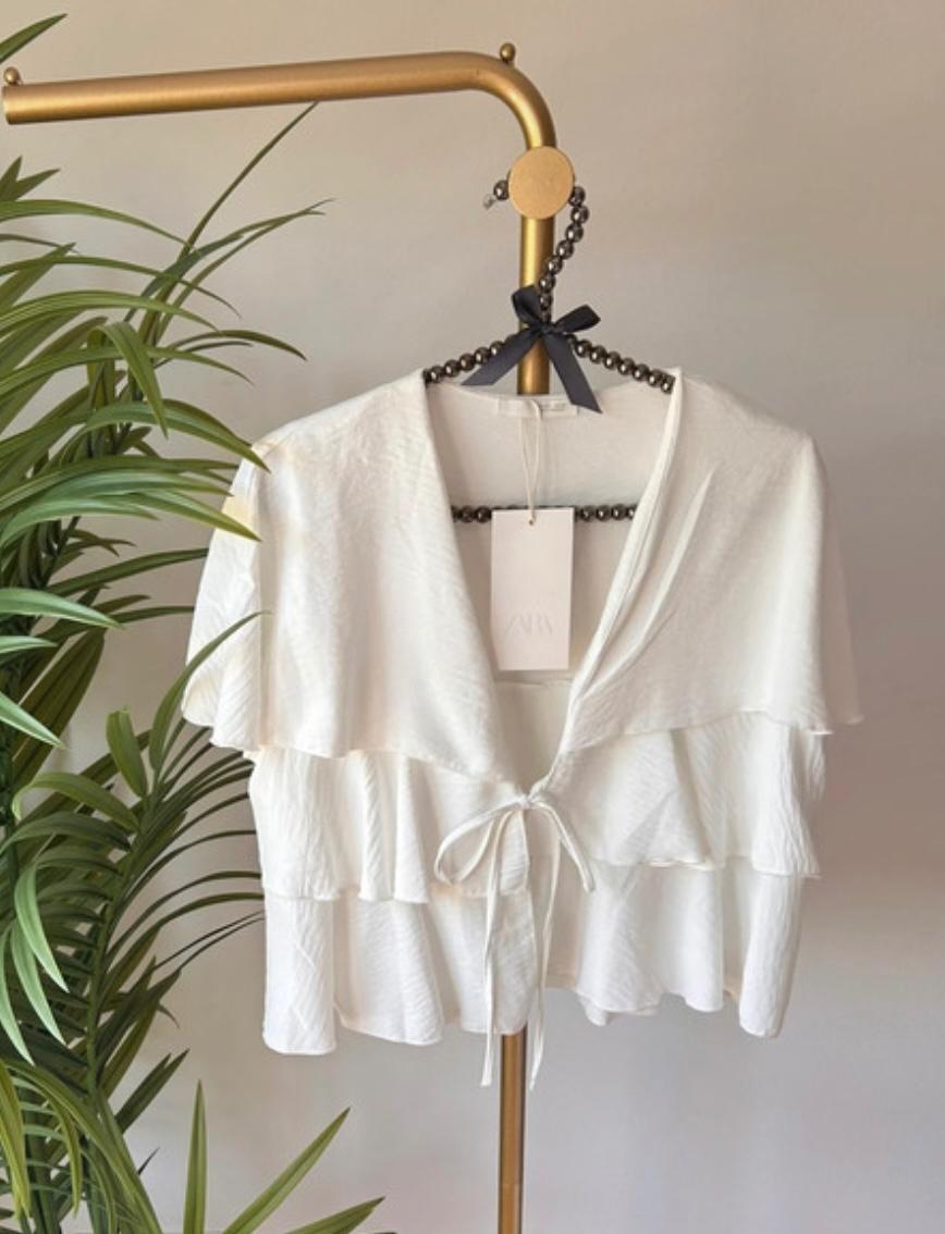 White top with ruffles and a front tie, hanging on a gold clothes rail next to a green plant. Label with the text ZARA.