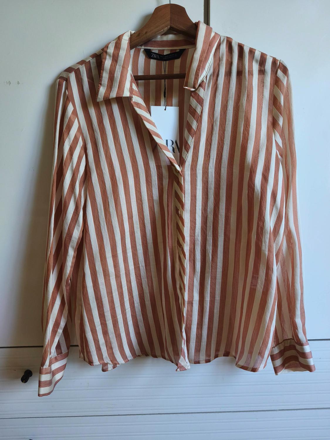 Long-sleeved shirt with white and orange vertical stripes hanging on a hanger. Label bearing the text ZARA.