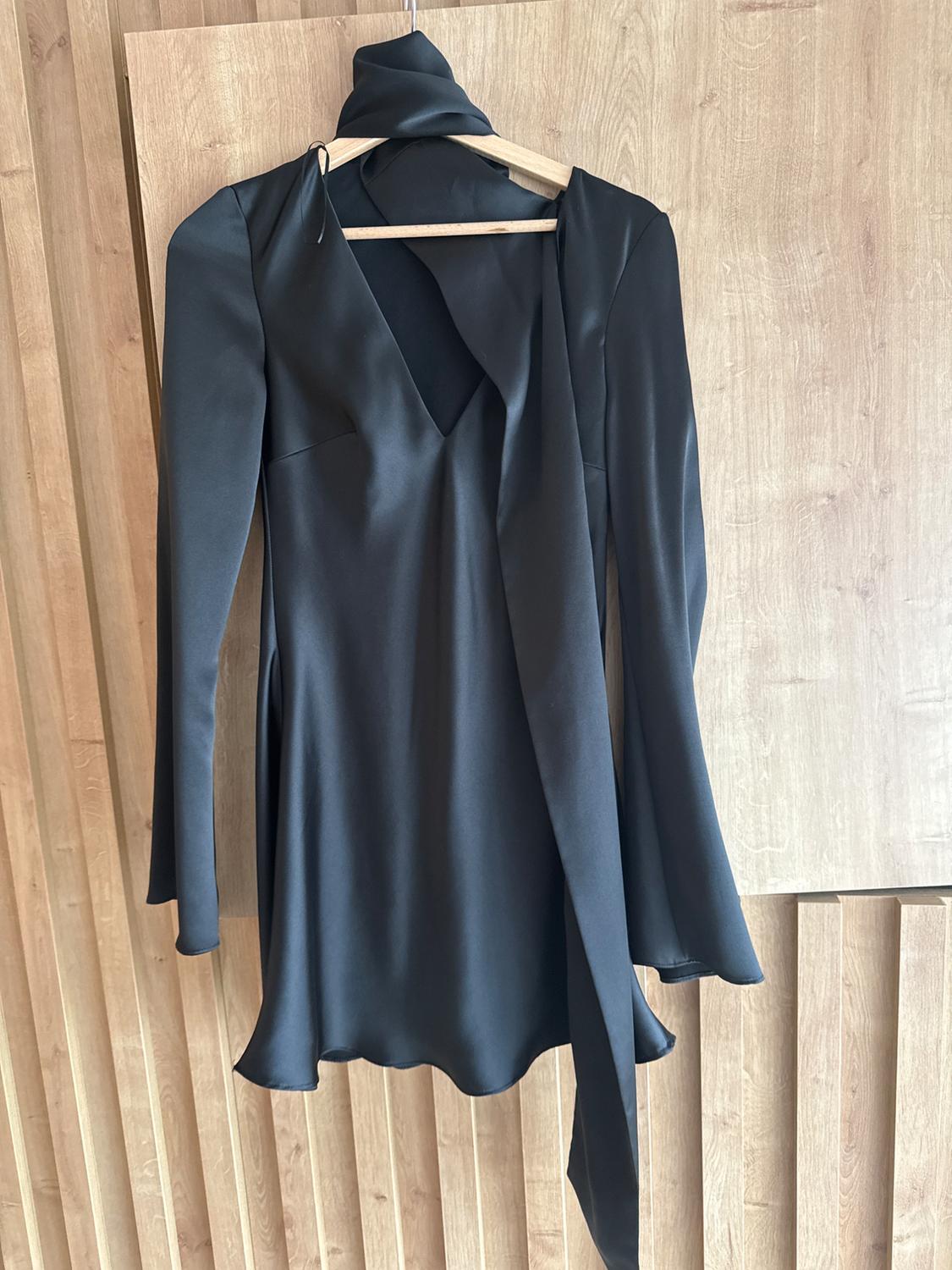 Black long-sleeved dress with a V-neck and a bow detail on the collar, hanging on a wooden hanger against a light wood background.