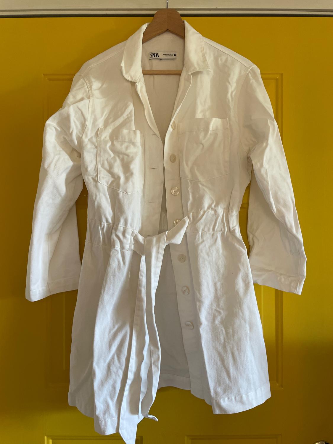 White coat with belt hanging on a hanger over a yellow door. Label visible on the collar.