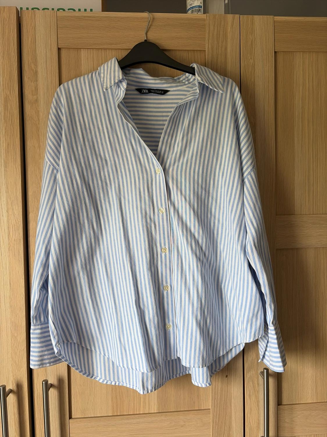 Blue and white striped shirt, tag with ZARA text, hanging on a black hanger in front of a light wood wardrobe. Relaxed atmosphere.