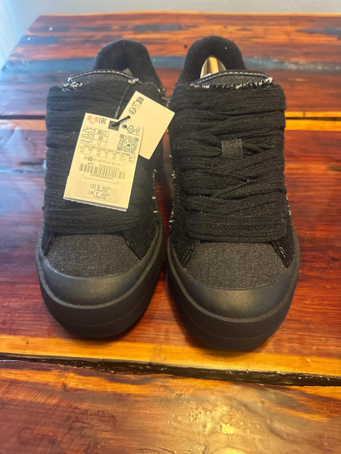 Black trainers with thick laces on a wooden table. Label: EAC ART: 2226/522/800 US $ 69.90 UK £ 49.99.