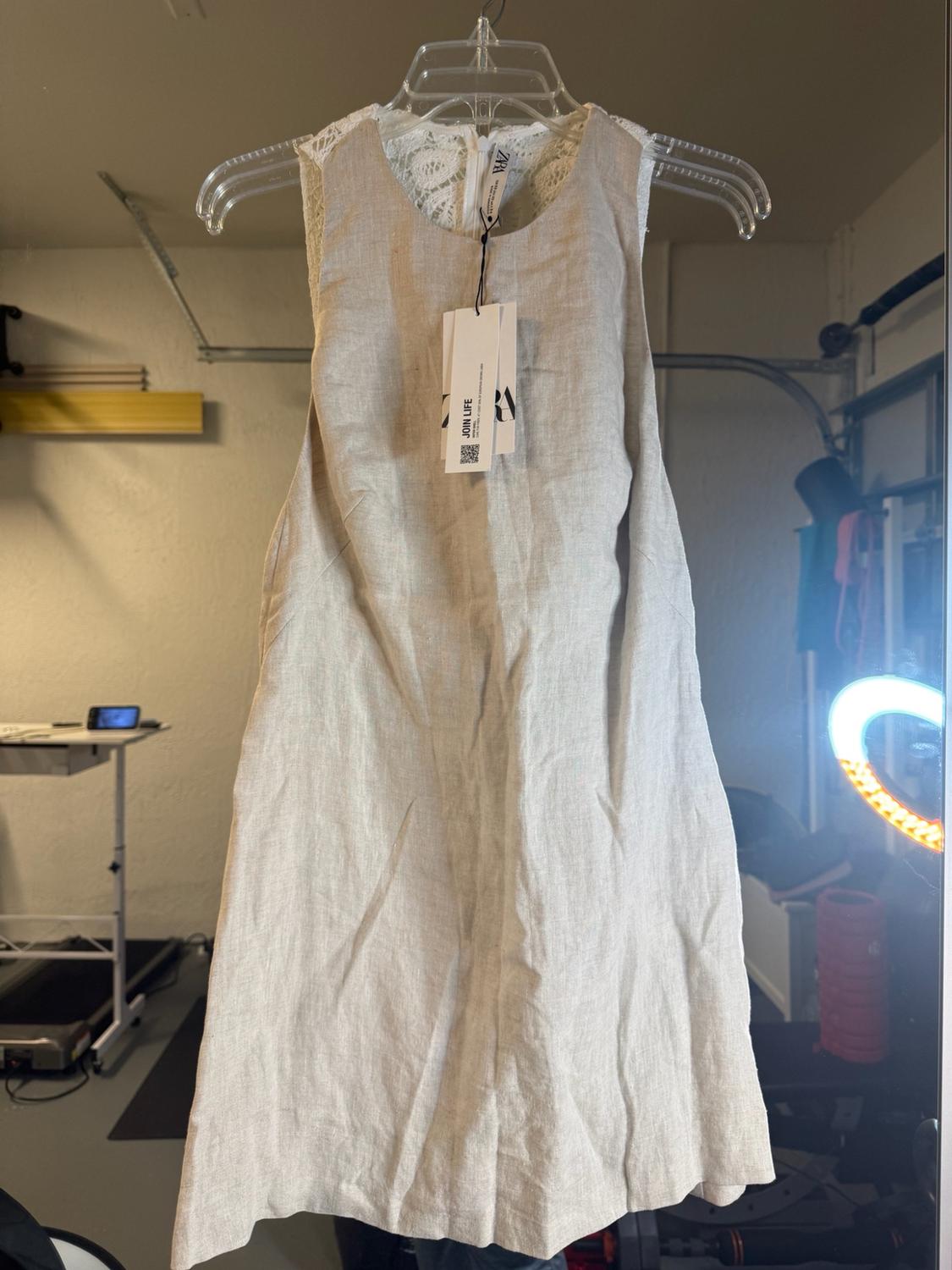 Beige sleeveless lace dress, hanging on a hanger in a room with a ring light. Labels with the text ZARA and JOIN LIFE.