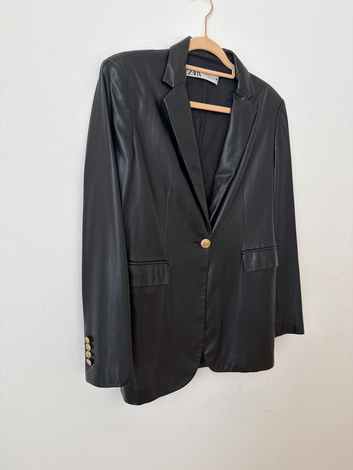 Black blazer hanging on a wooden hanger against a white wall. It features gold buttons, flap pockets and a ZARA label.