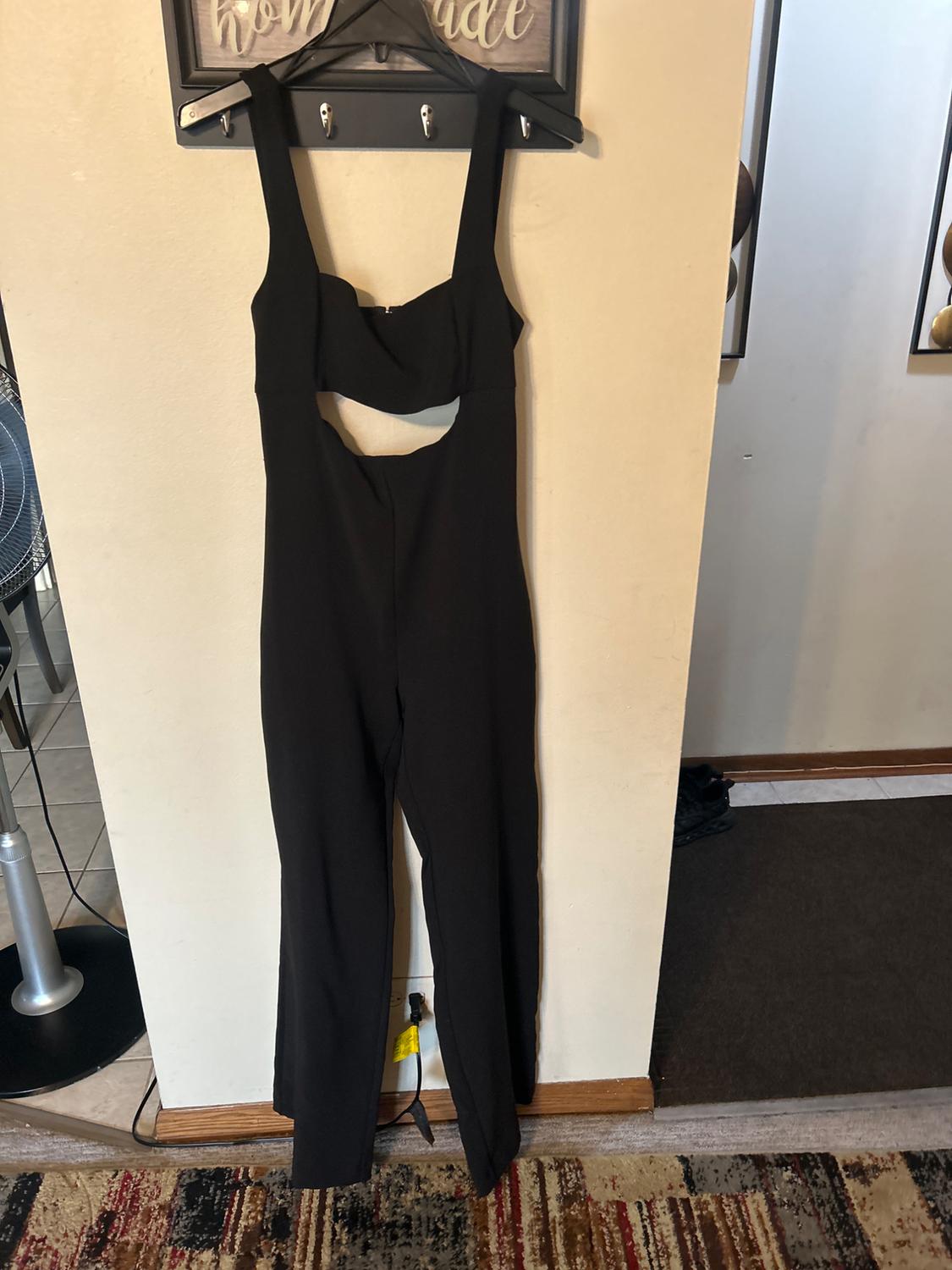 Black jumpsuit with cut-out neckline hanging on a hanger against a beige wall. Colourful rug and fan in the background. Text: "homemade".
