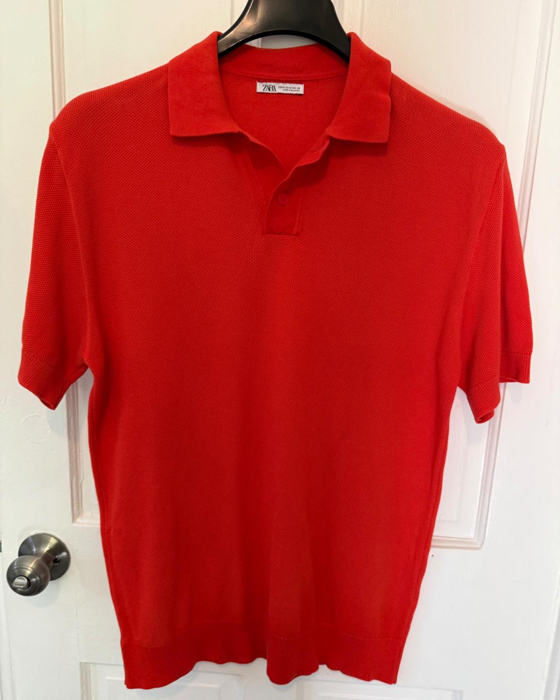 Red short-sleeved polo shirt hanging on a black hanger over a white door. Visible tag on the collar.