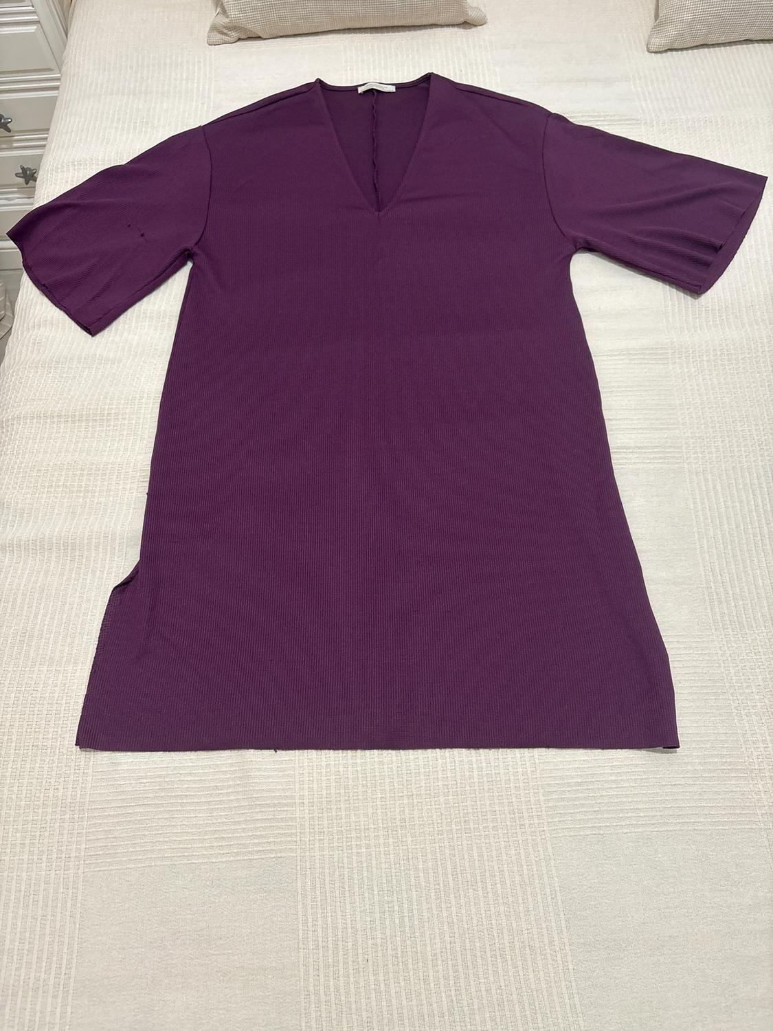 Purple short-sleeved V-neck t-shirt, spread out on a bed with a white bedspread and beige cushions.