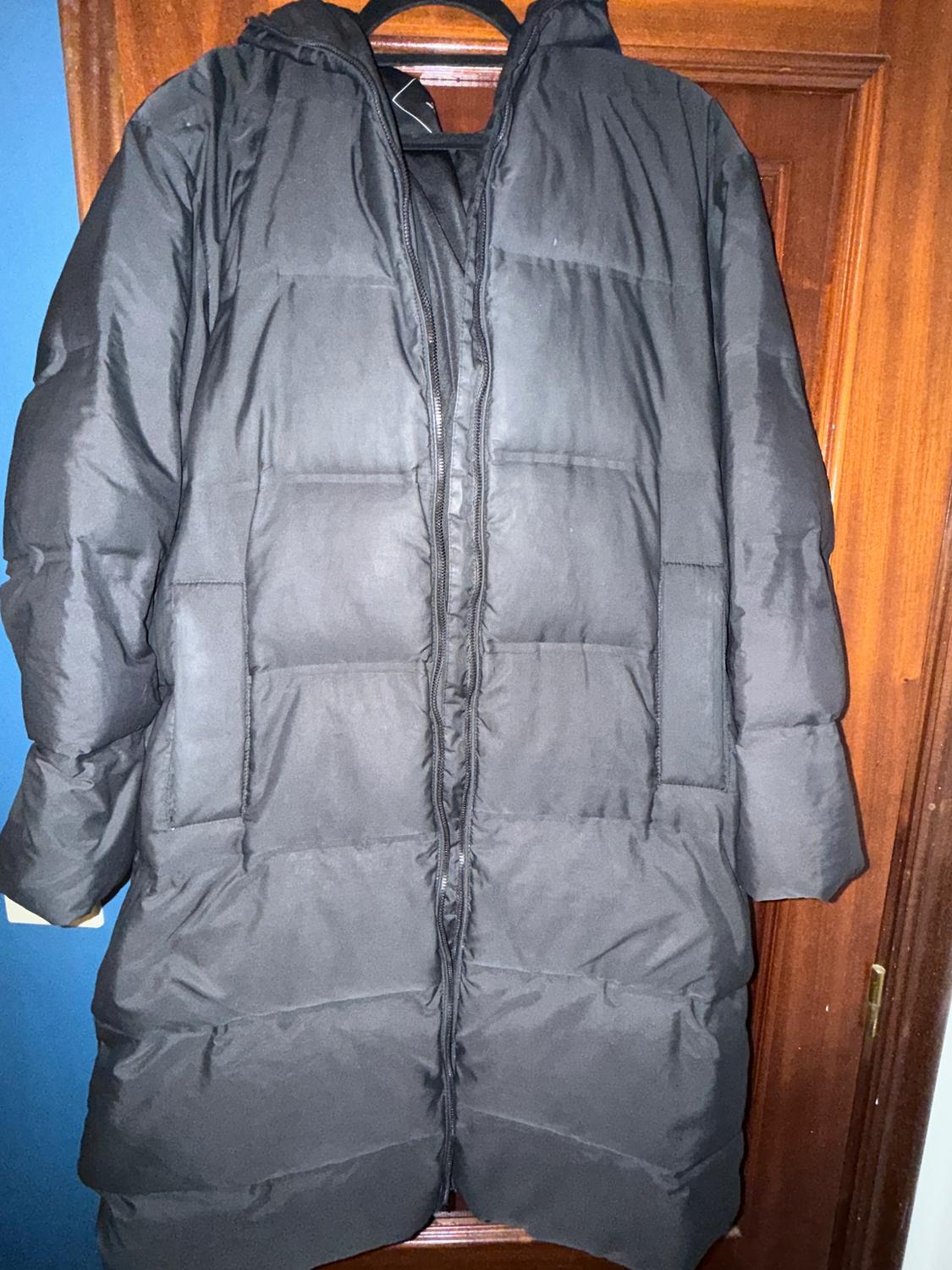 Black padded coat hanging on a wooden door. The coat has a hood and a front zip fastening.