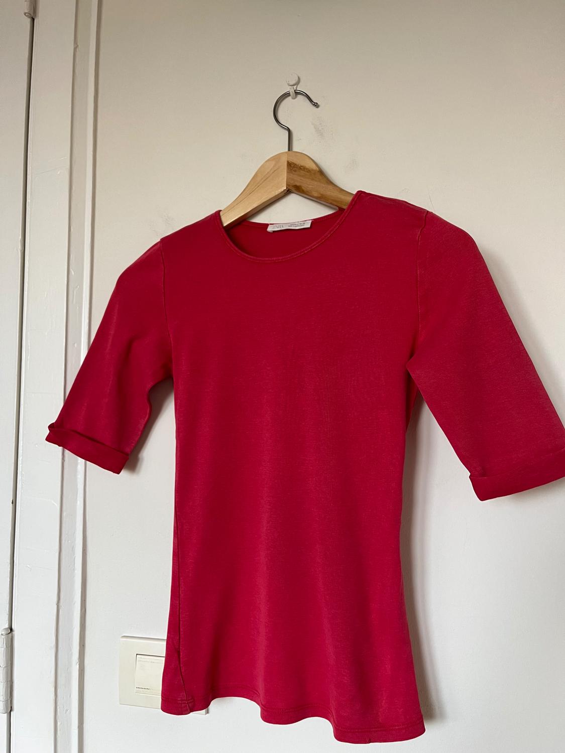 Red three-quarter sleeve t-shirt hanging on a wooden hanger against a white wall.