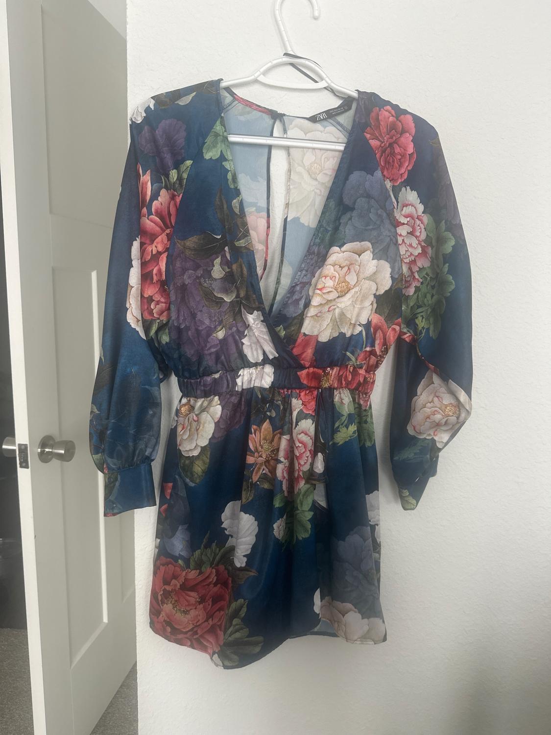 Short dress with a floral print in red, green, and blue tones, hanging on a white hanger against a white wall.