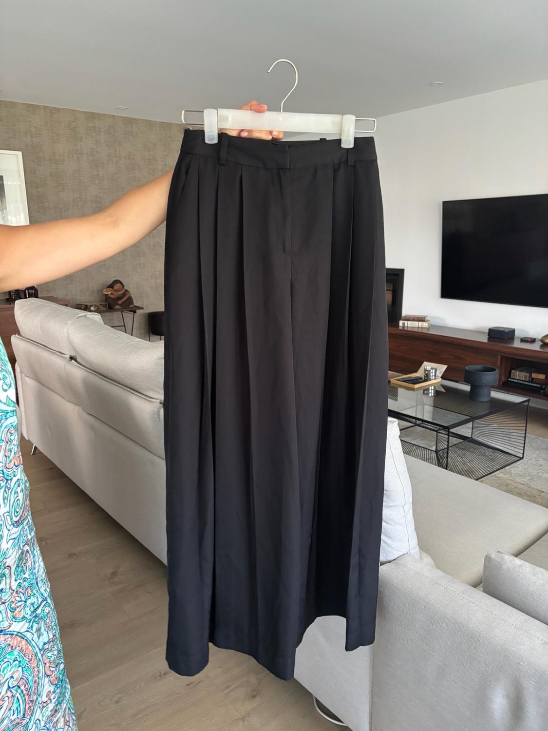 Arm holding a pair of wide-leg black pleated trousers on a white hanger, in a modern living room with a light grey sofa and television in the background.