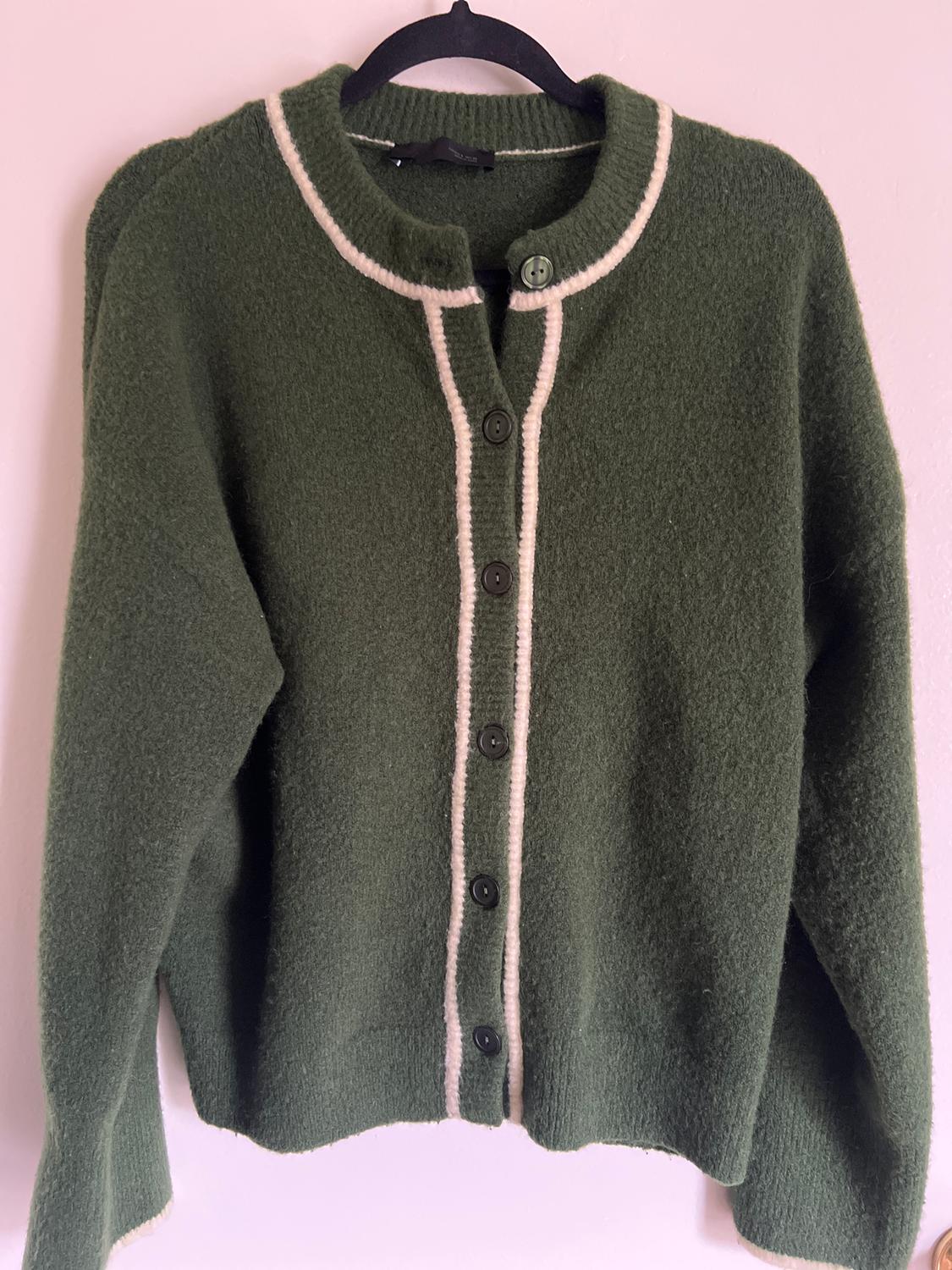 Green wool cardigan with white trim and black buttons, hanging on a black hanger against a white background.