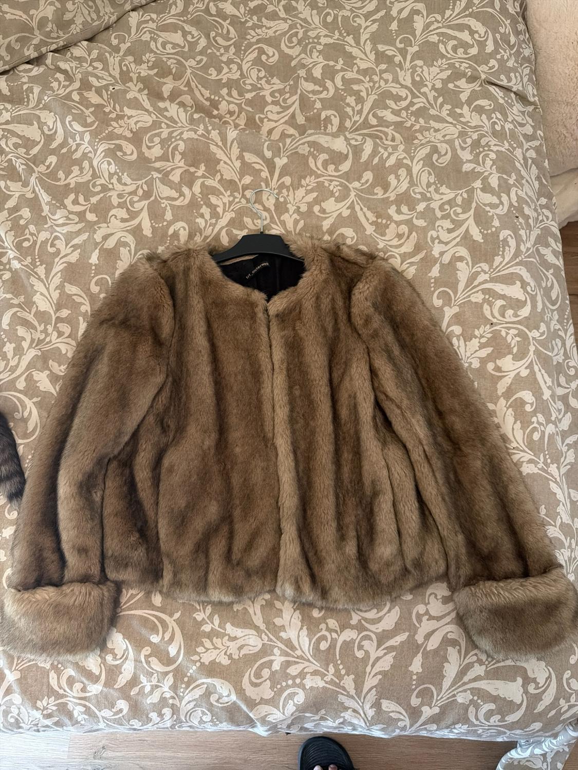 Short brown fur jacket on a hanger, laid out on a bed with a beige floral-patterned bedspread.